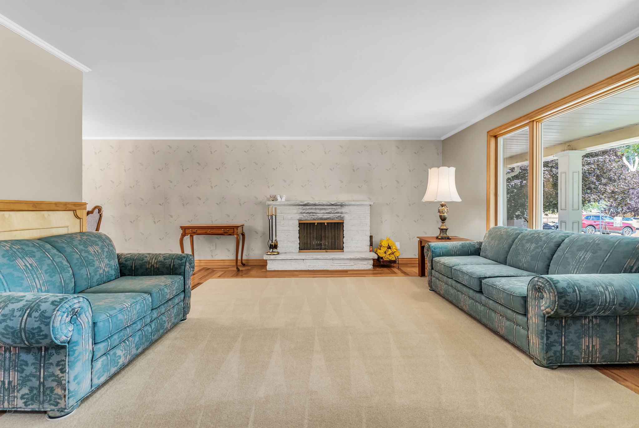 Living space featuring a white brick fireplace with a metal grate, natural wood-finish flooring bordering a light-toned carpet, and a large window with wood trim