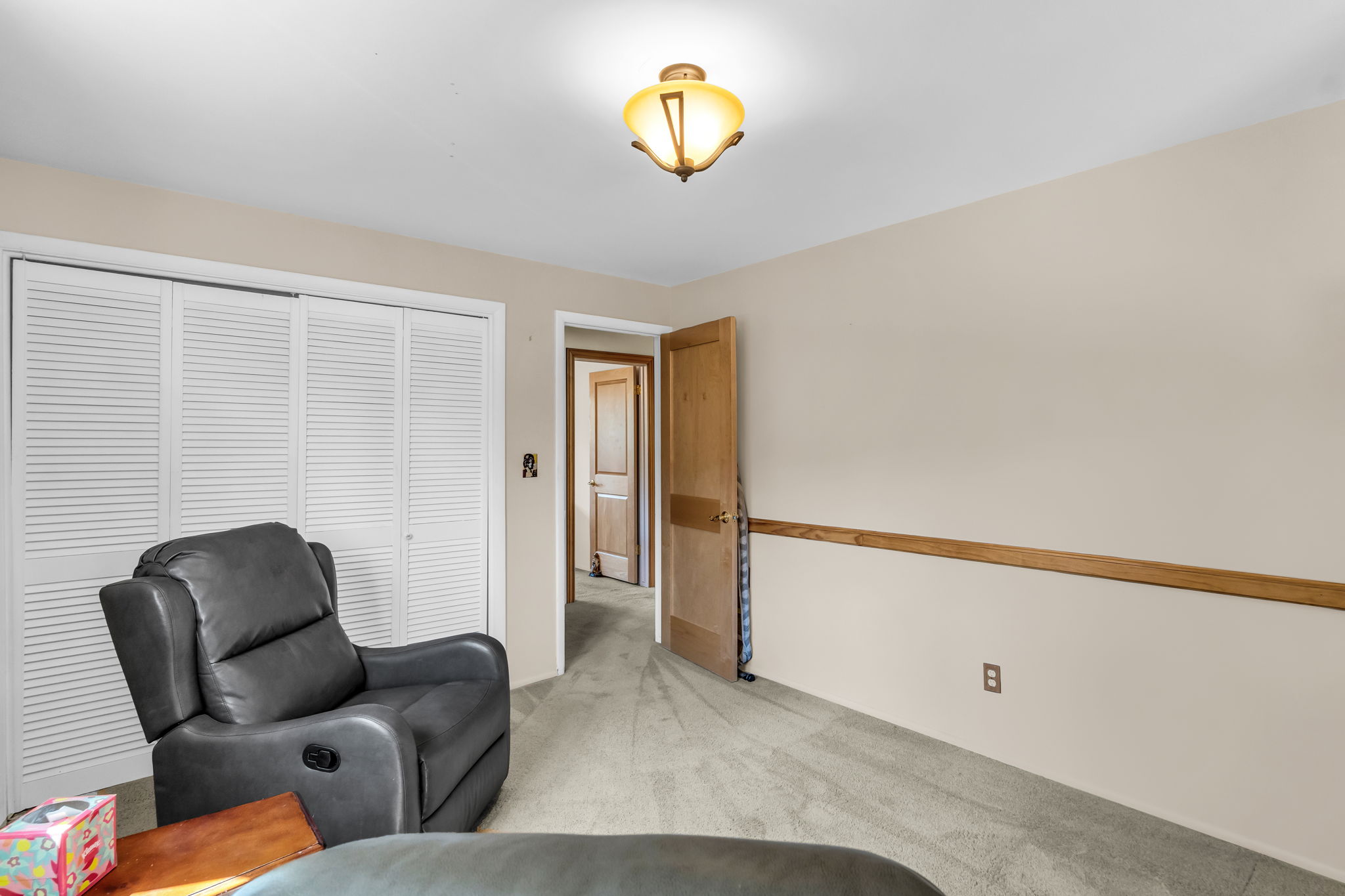 Neutral wall paint, light-colored carpeting, white louvered bifold closet doors, natural wood trim, and a semi-flush mount ceiling light fixture characterize this room