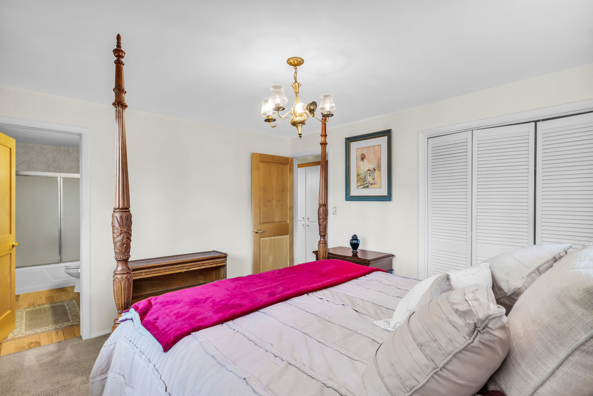 Carpeted bedroom featuring a brass chandelier, louvered closet doors, a natural wood door, and an entryway to a bathroom with a white bathtub