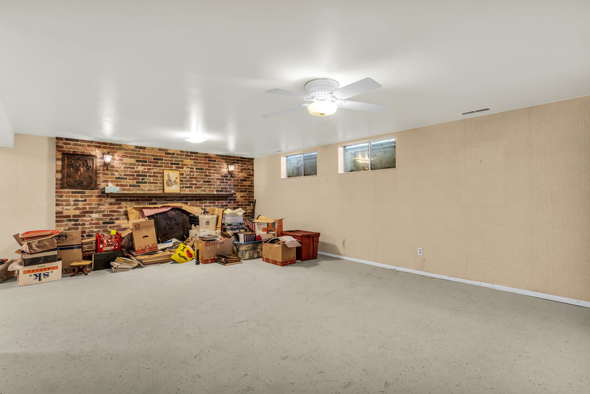 Spacious lower-level room featuring a brick accent wall with a wood mantel, recessed lighting, and a ceiling fan