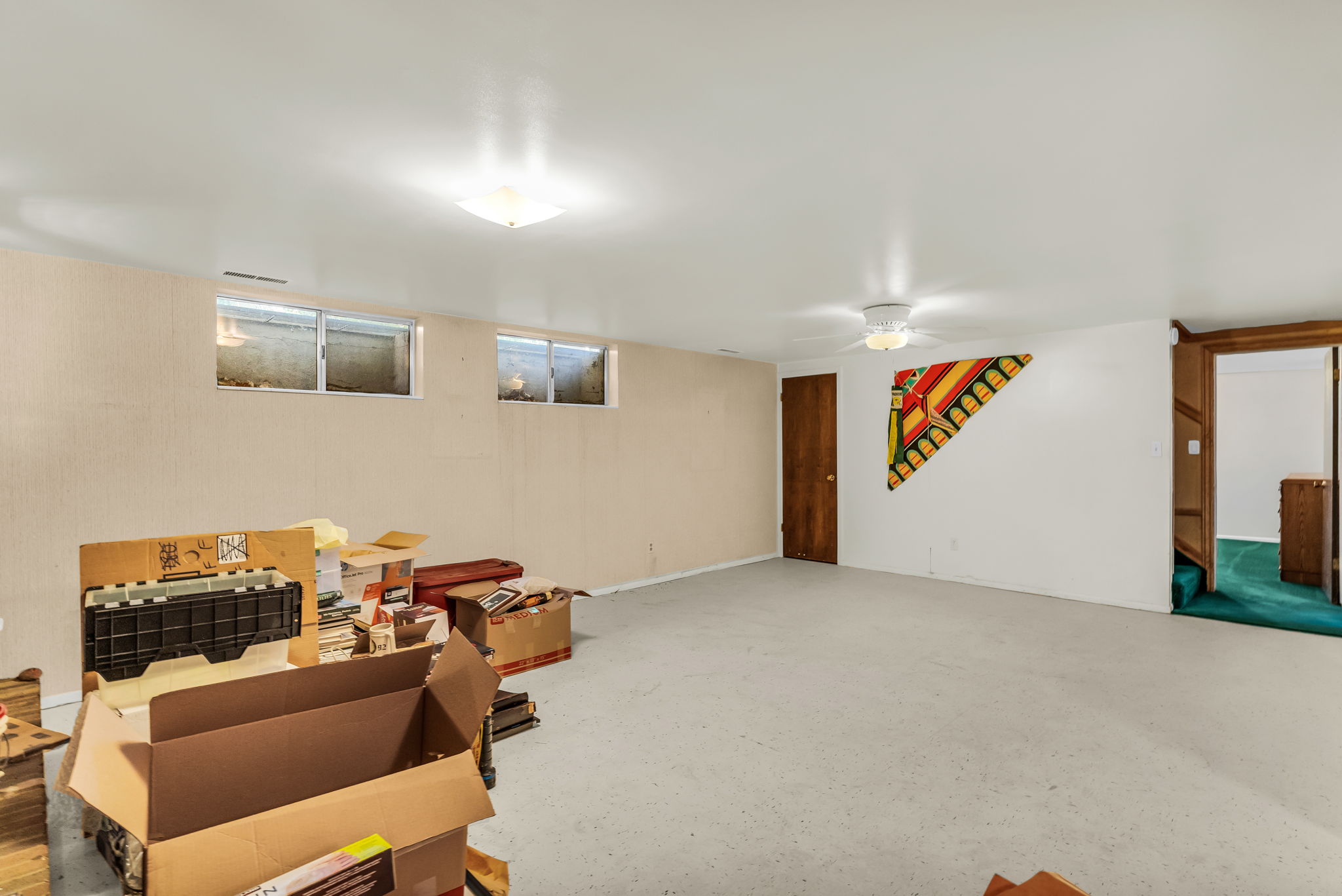 Spacious lower-level room featuring light-toned walls, recessed windows, an overhead light fixture, a ceiling fan, and a wood-paneled doorway