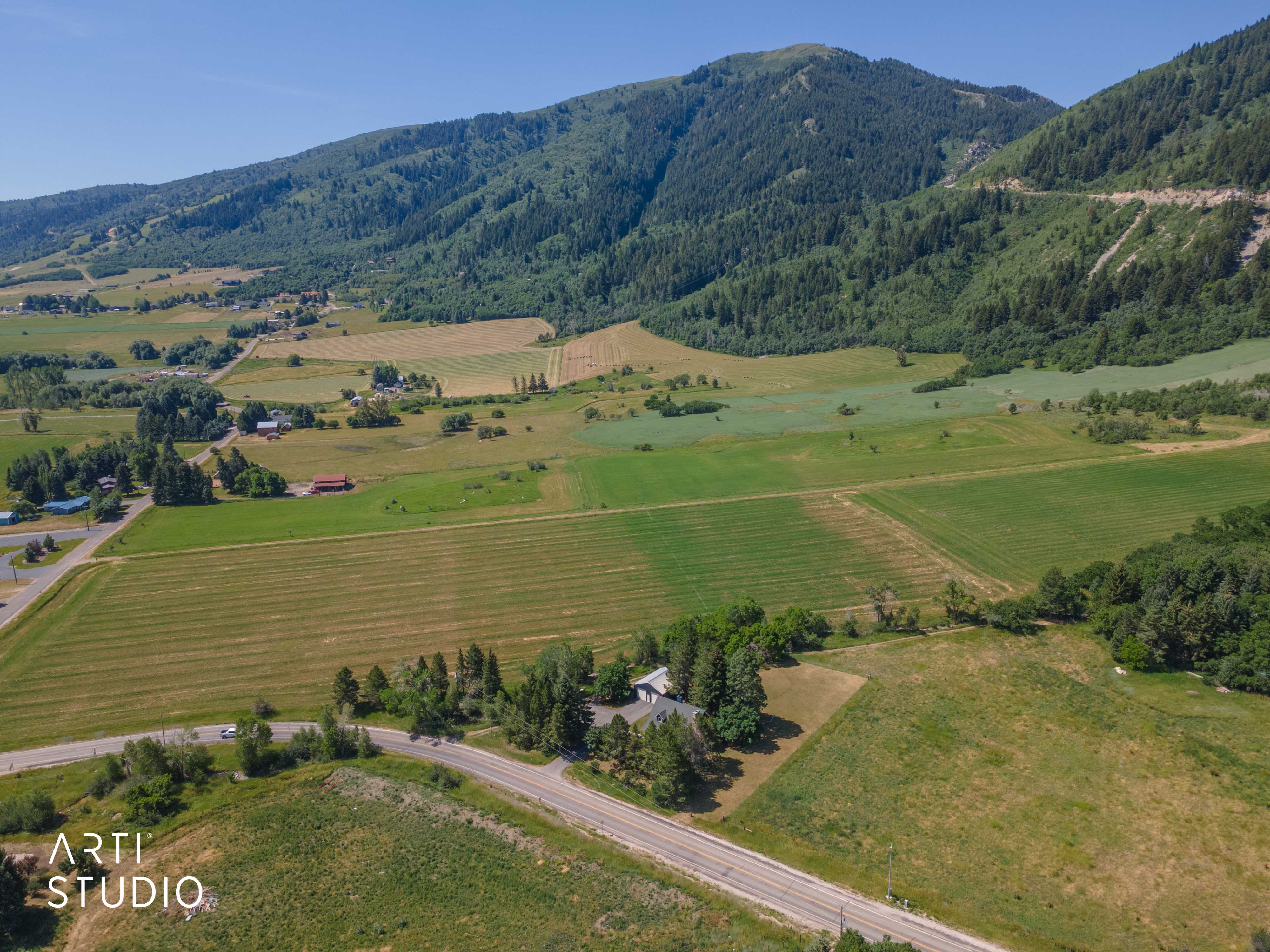 Expansive property featuring verdant fields, a primary residence with an attached garage, a paved road frontage, and a backdrop of verdant mountains