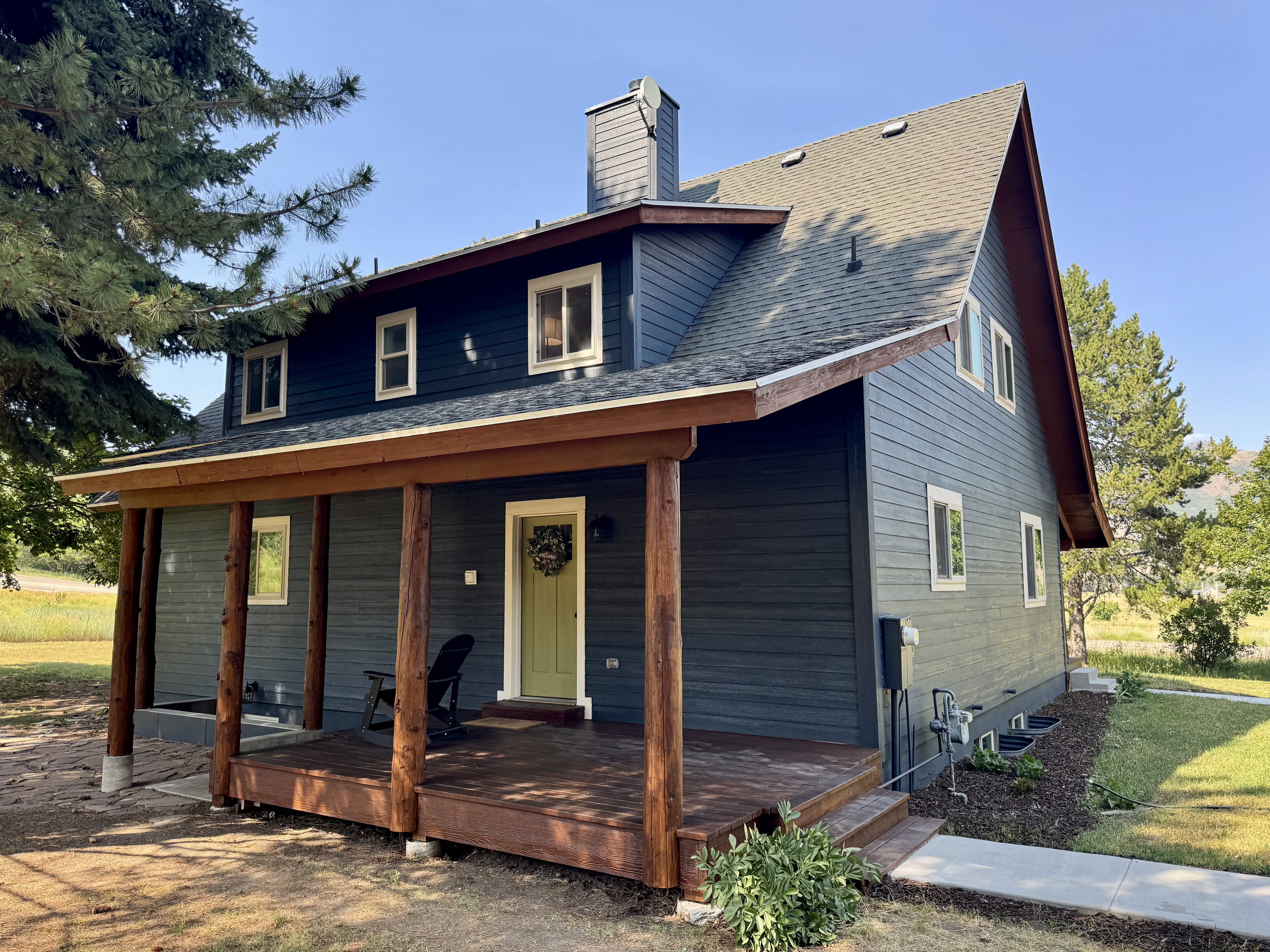 Residential exterior featuring dark blue siding, a covered porch with natural wood posts and decking, a light green entry door, a prominent chimney, and multiple windows with white trim