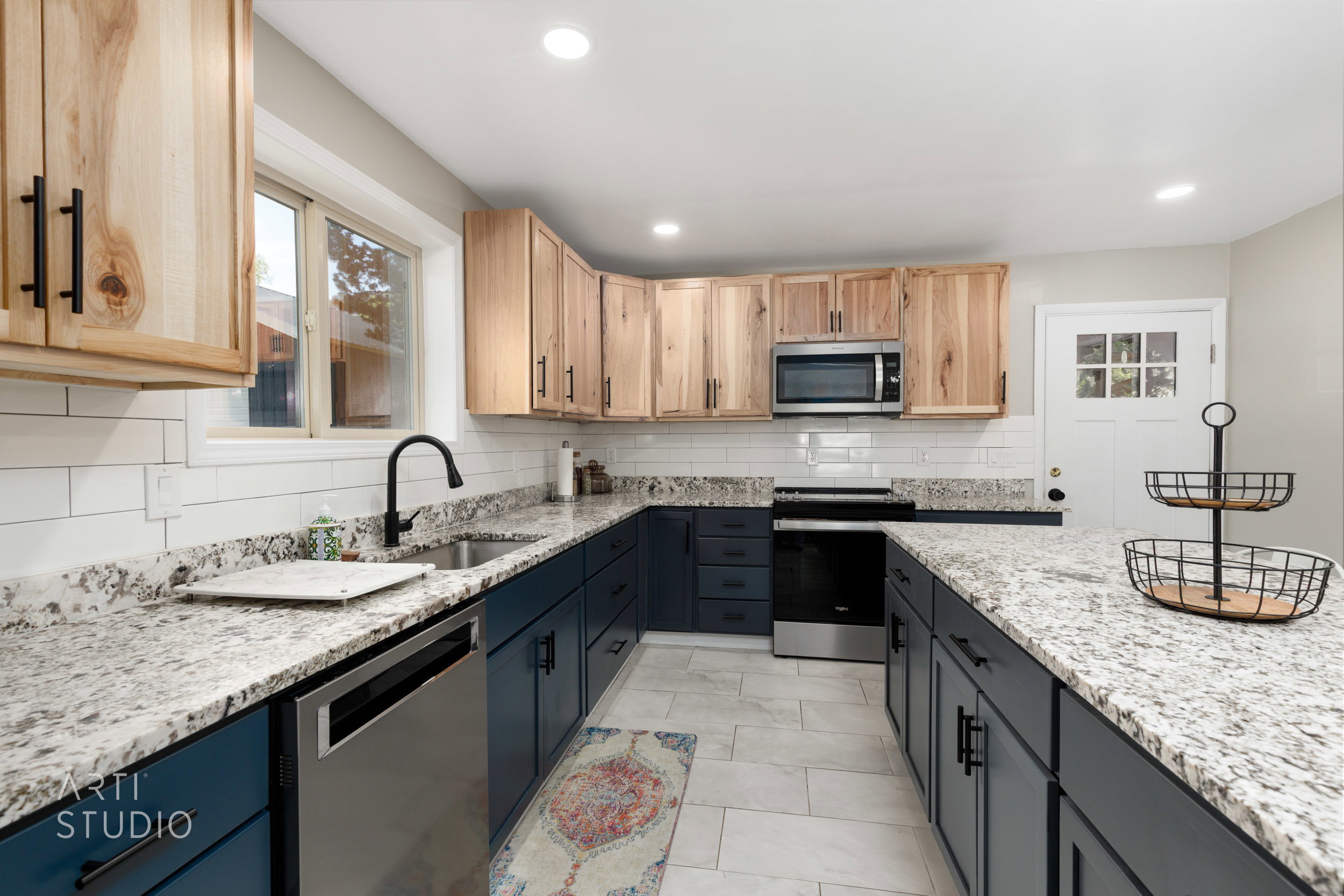 Contemporary kitchen featuring two-tone cabinetry with natural wood-finish upper cabinets and matte blue lower cabinets, granite countertops, subway tile backsplash, and stainless steel appliances