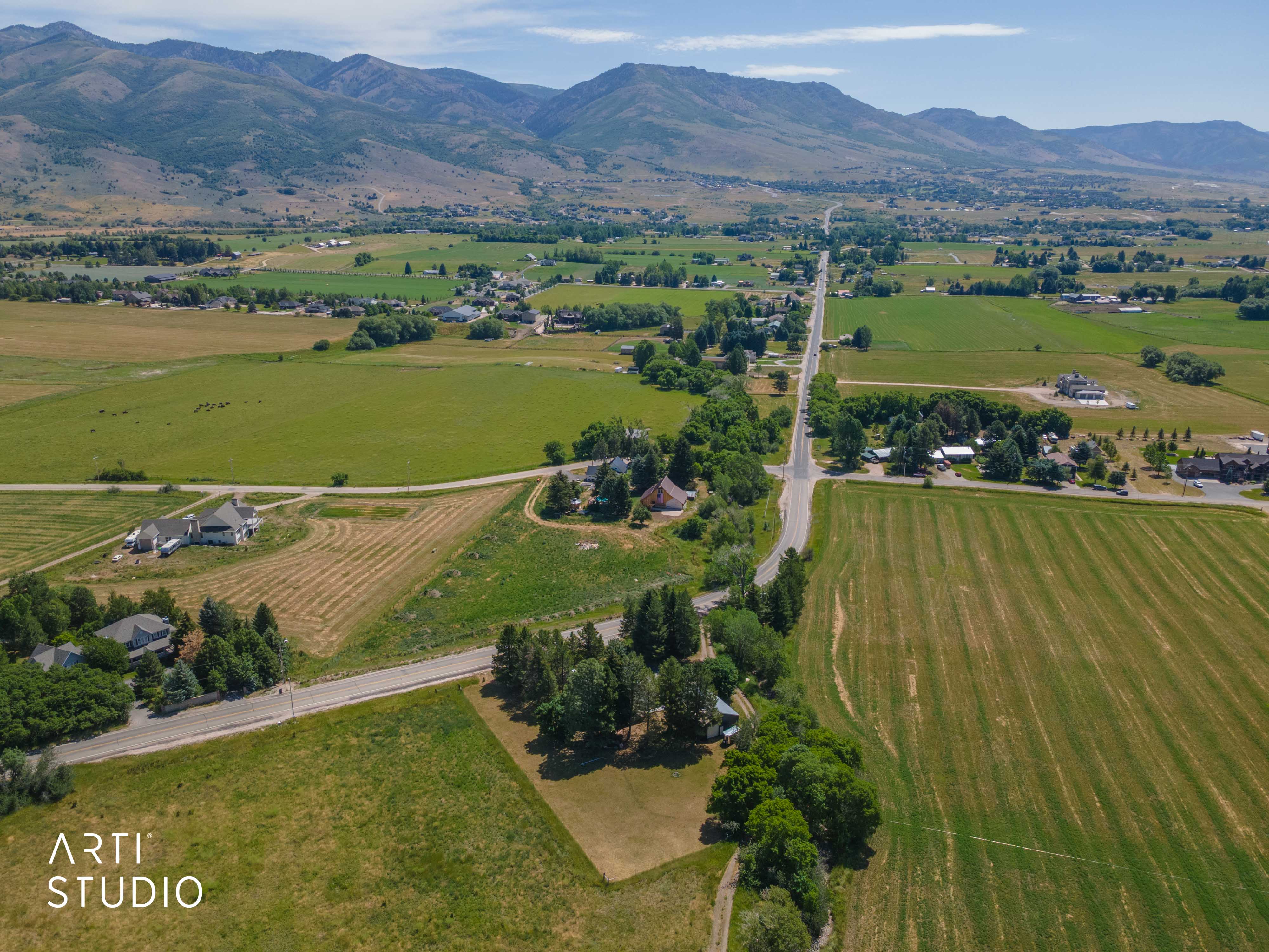 Expansive rural landscape featuring a paved road, scattered residential properties, mature trees, open fields, and distant mountains