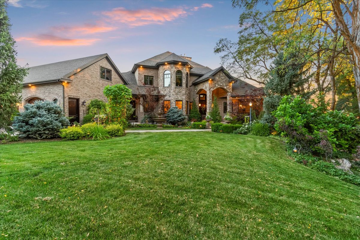 Grand exterior featuring a stone and brick facade, arched entryway, multi-paned windows, a manicured lawn, and mature landscaping