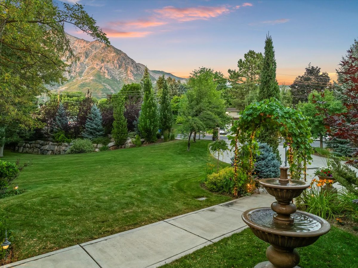 Expansive front yard featuring a tiered water fountain, mature landscaping, concrete pathway, decorative archway, and mountain backdrop