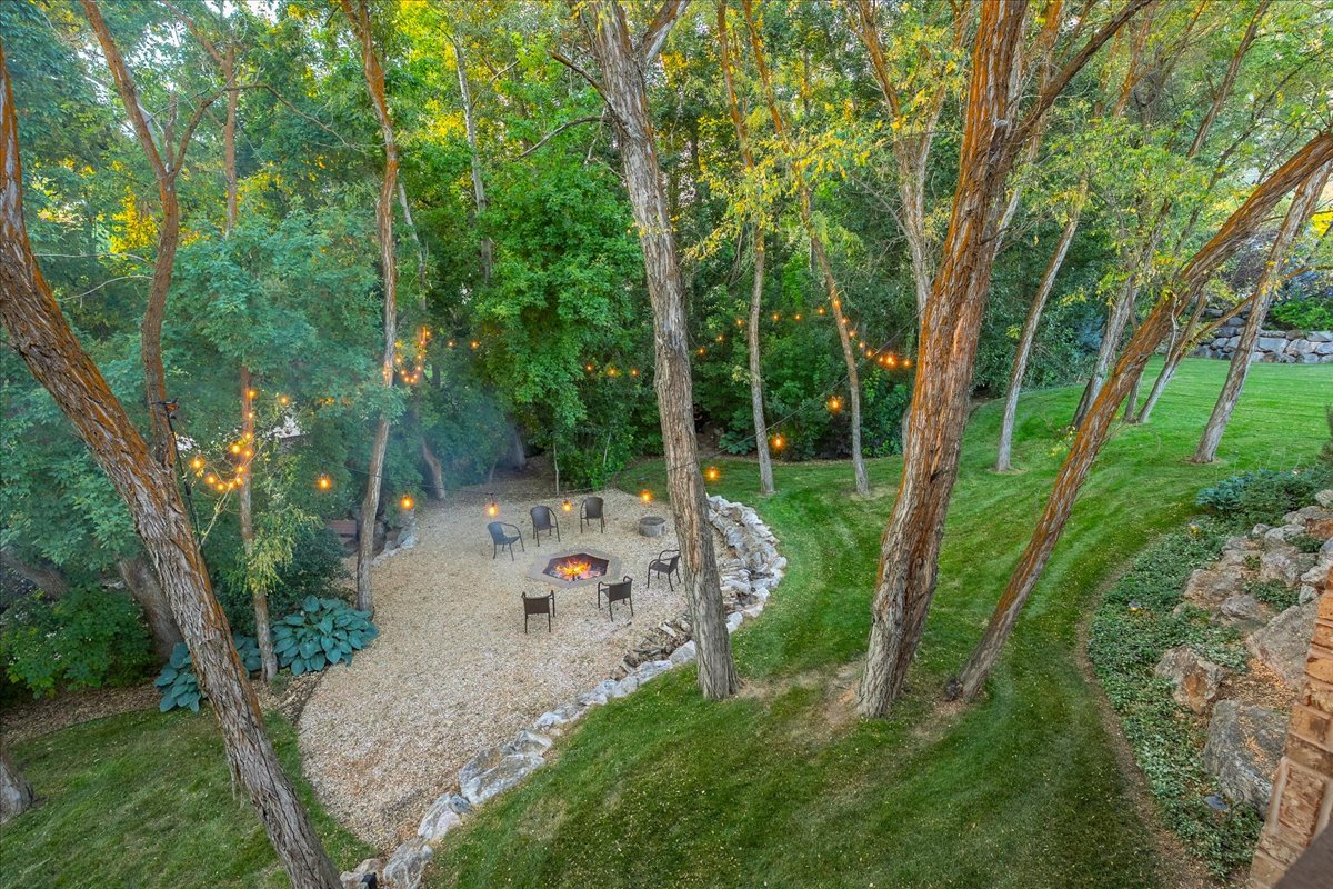 Expansive outdoor entertaining area featuring a gravel fire pit with built-in seating, surrounded by mature trees, string lighting, and manicured lawns