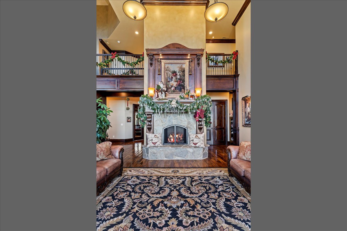 Great room featuring a two-story stone fireplace with an ornate wood mantel, hardwood flooring, and an upper-level wood railing