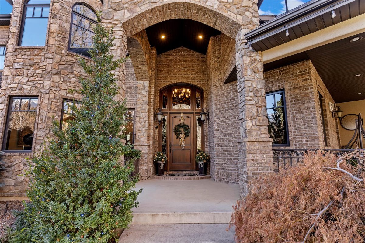 Stone and brick exterior featuring an arched entryway, dark wood front door with glass panels, and a covered porch with a metal railing