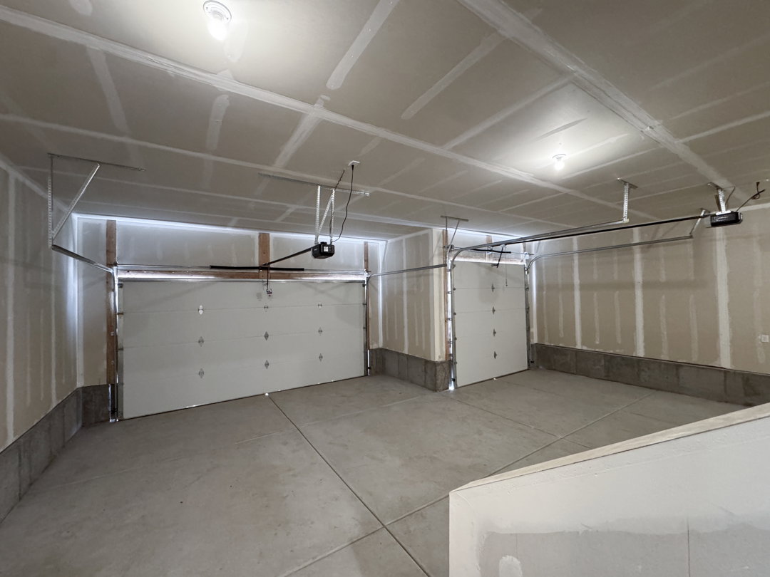 Spacious two-car garage featuring two separate overhead doors, exposed drywall, concrete flooring, and ceiling-mounted lighting fixtures