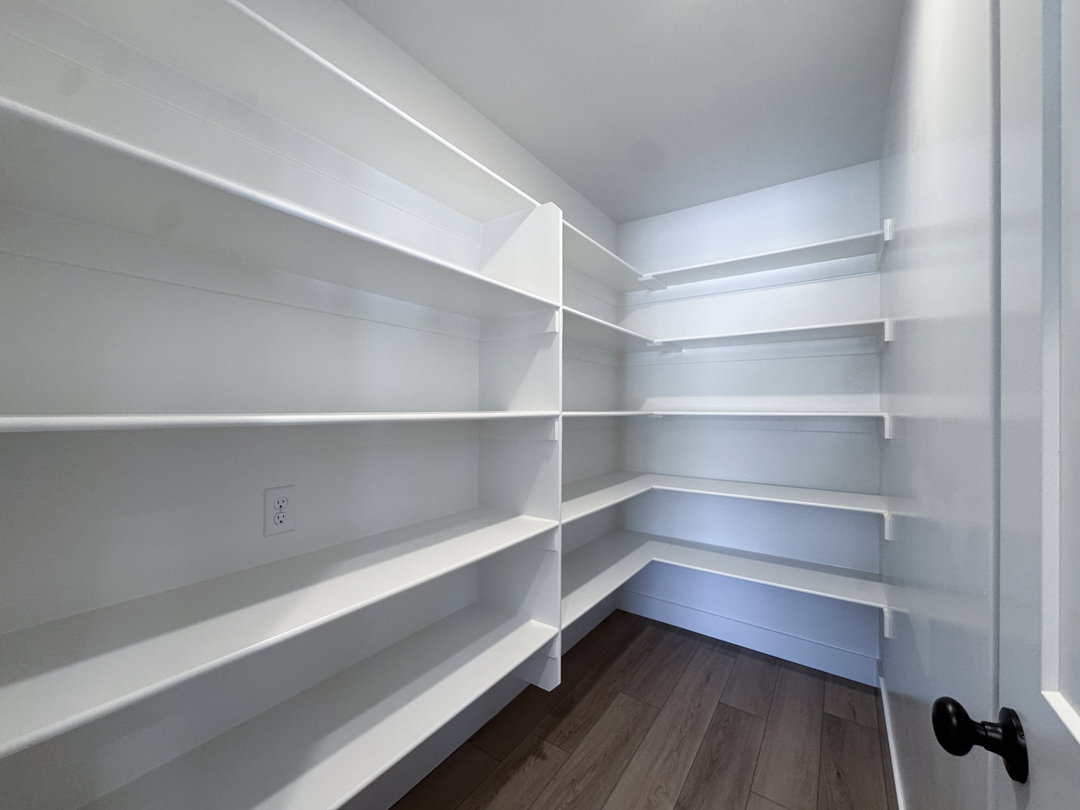 Spacious walk-in pantry featuring extensive white shelving, wood-finish flooring, and a door with a matte black knob