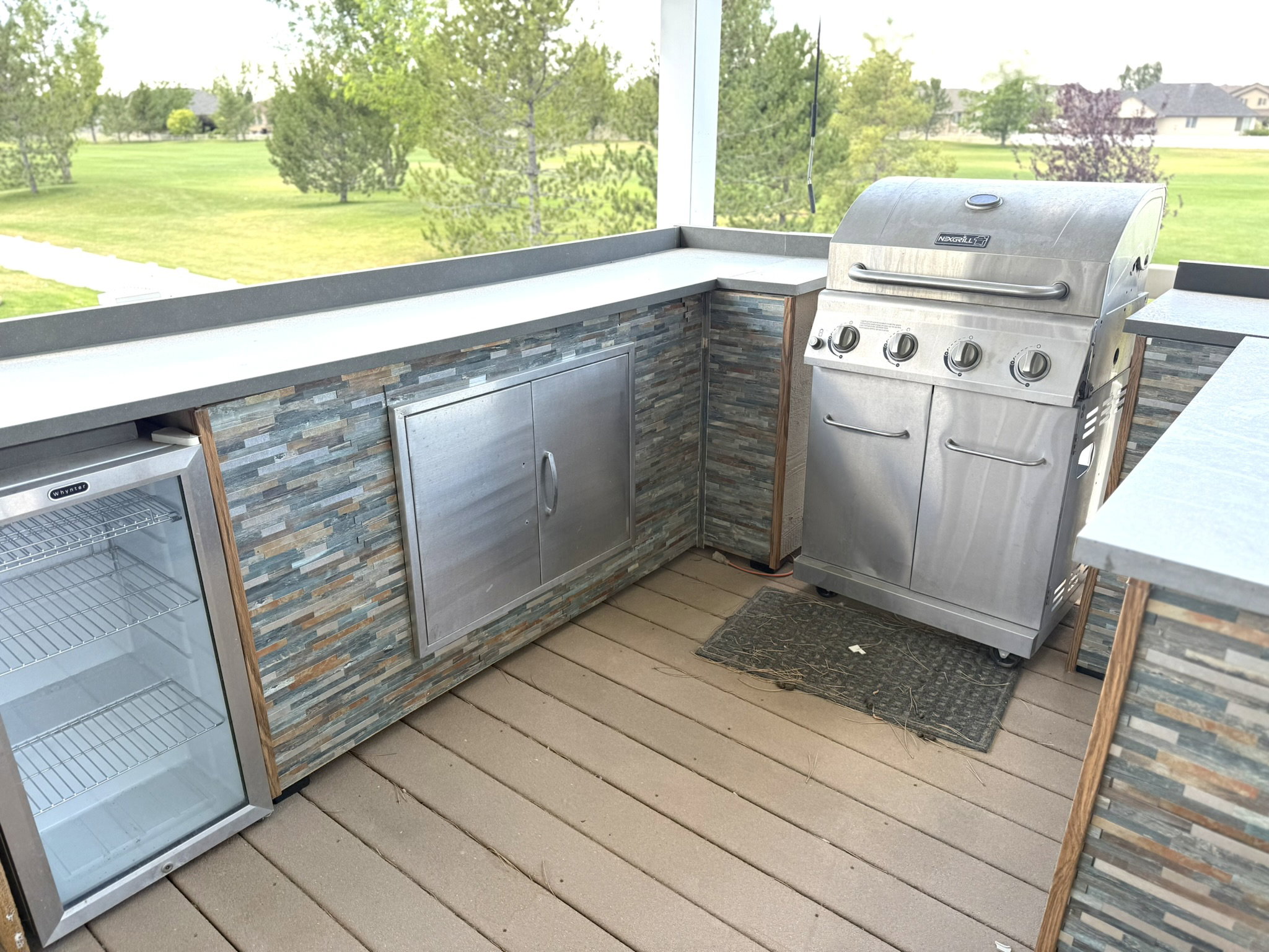 Outdoor kitchen featuring a built-in stainless steel grill, integrated under-counter refrigerator, ample counter space, and stacked stone veneer accents