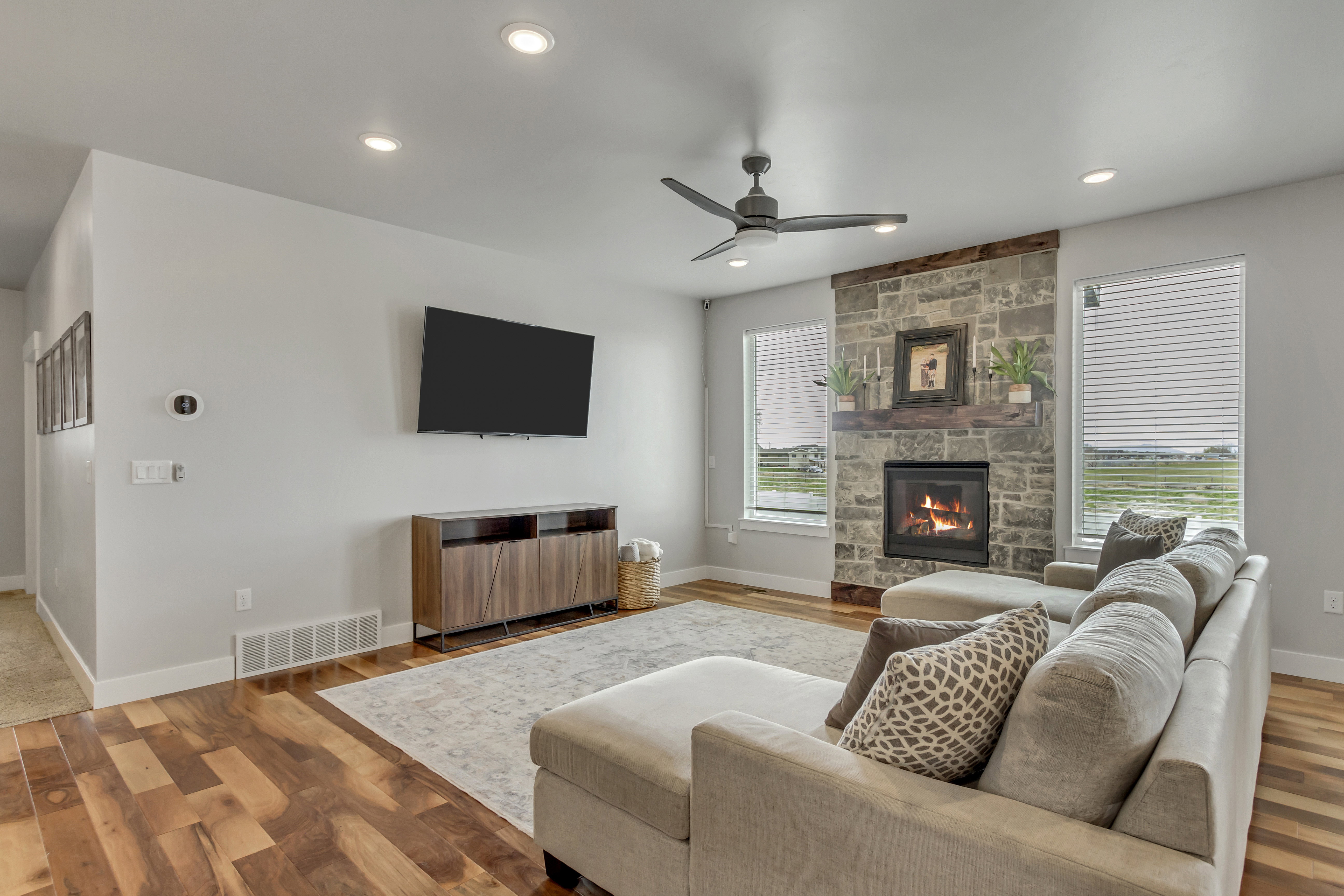Living area featuring a stone-clad fireplace with a wood mantel, wood-finish flooring, recessed lighting, a ceiling fan, and multiple windows with blinds