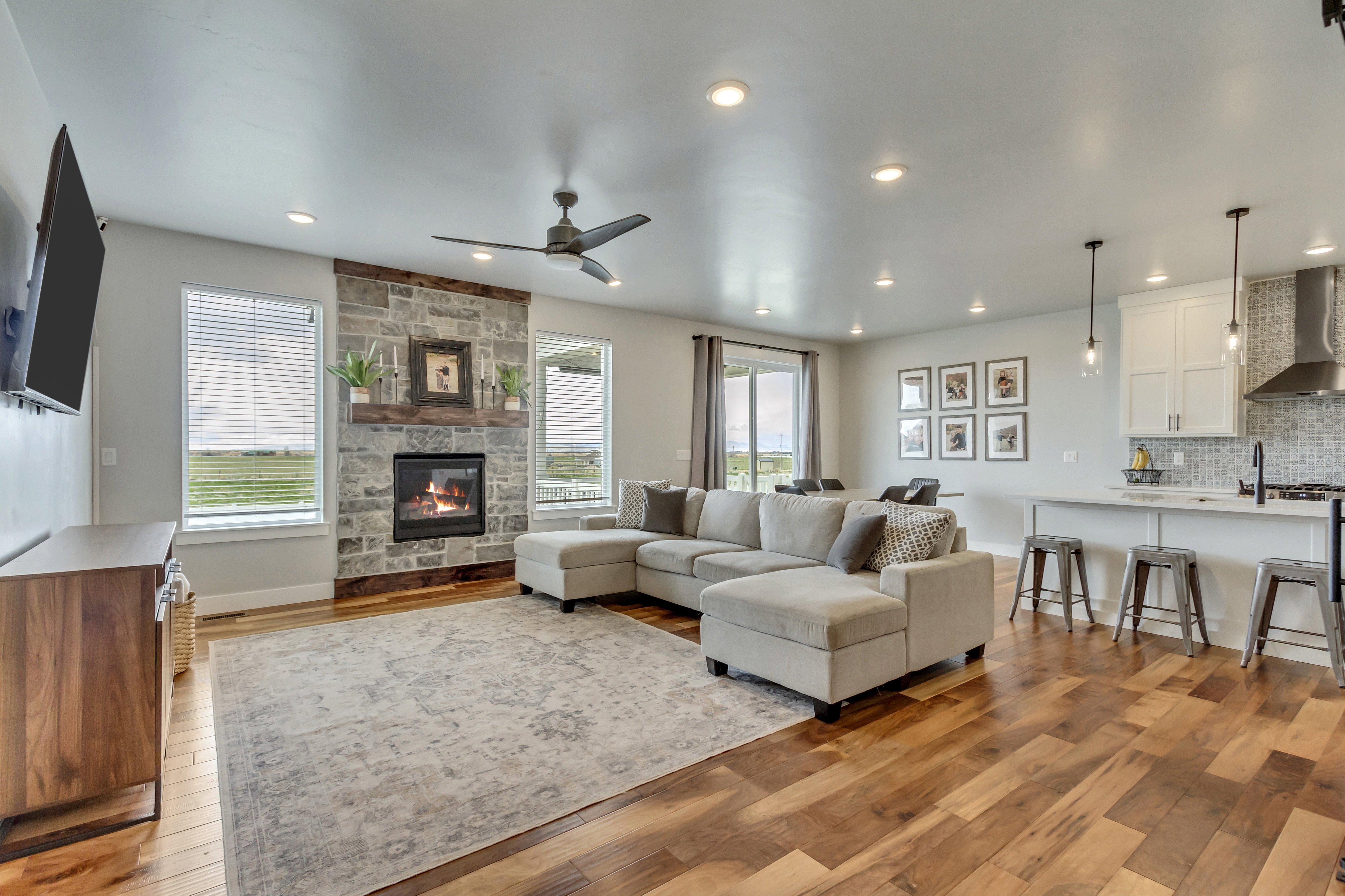 Open-concept living area featuring wood-finish flooring, a stone-faced fireplace with a wood mantle, recessed lighting, and a contemporary ceiling fan