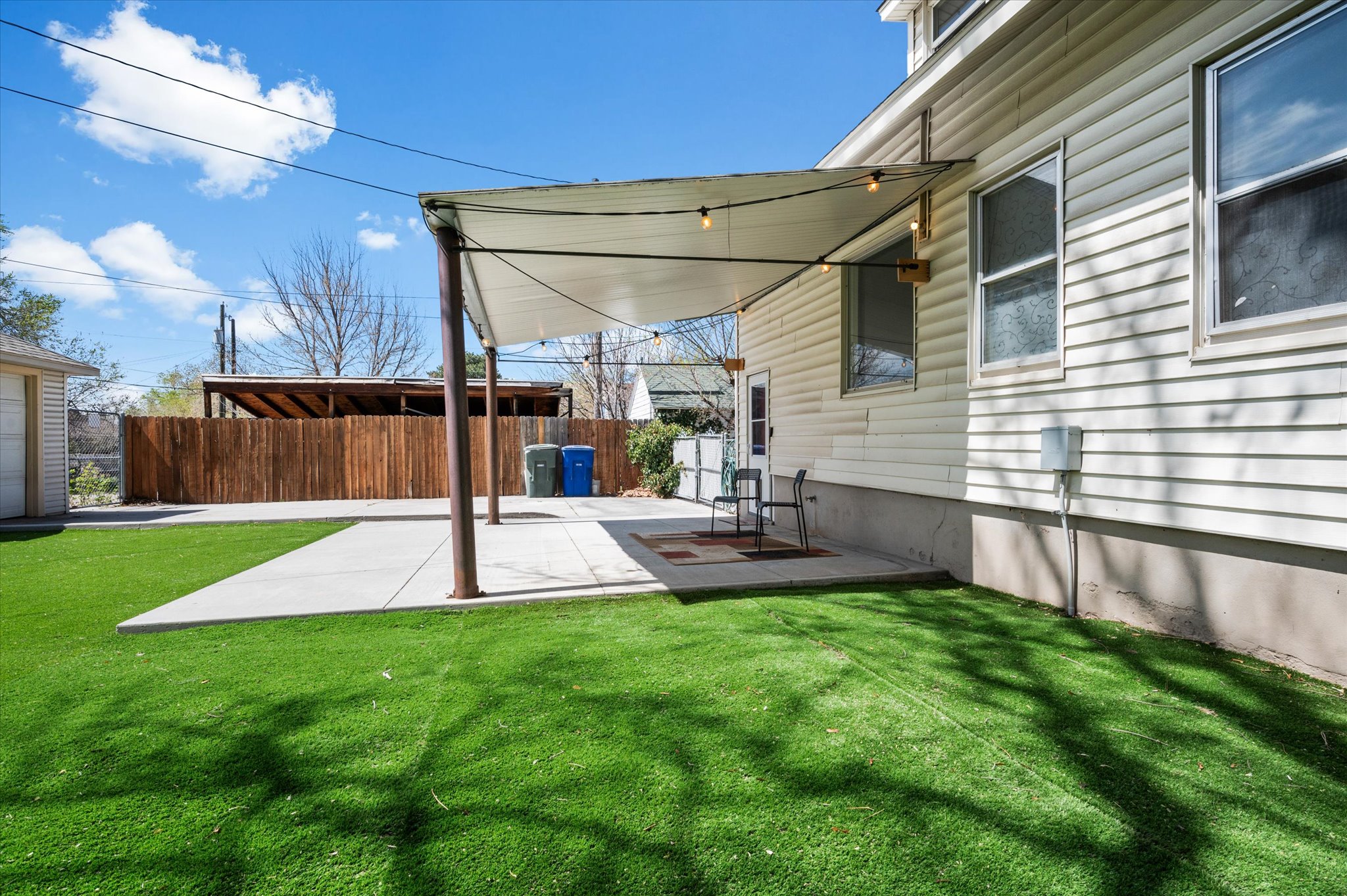 Patio with overhead shade canopy and string lighting, complemented by a concrete patio pad, artificial turf, and a wood privacy fence