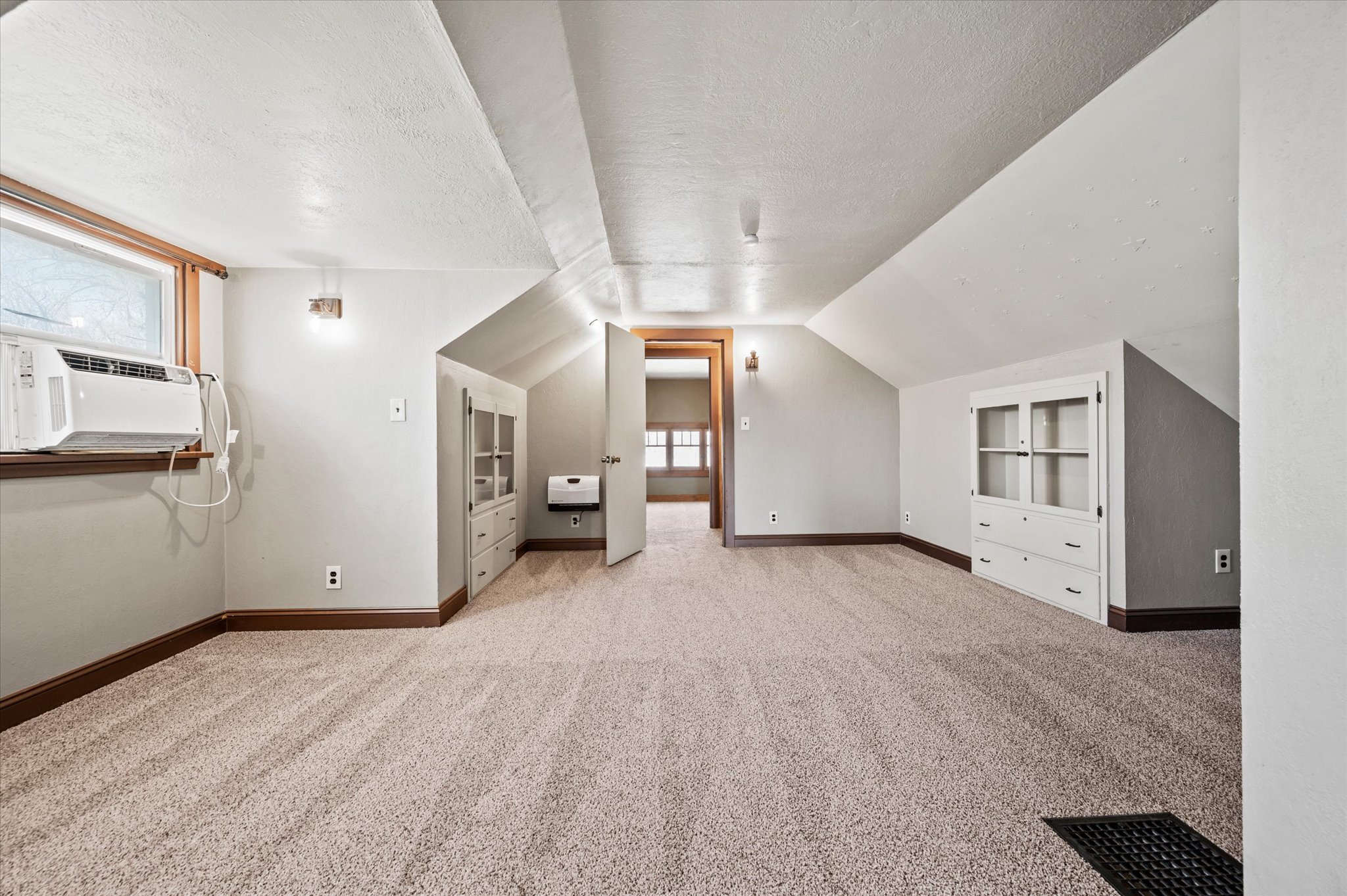 Carpeted room with sloped ceilings, featuring a built-in window unit, wall sconces, and built-in cabinetry with glass-front uppers and drawer storage