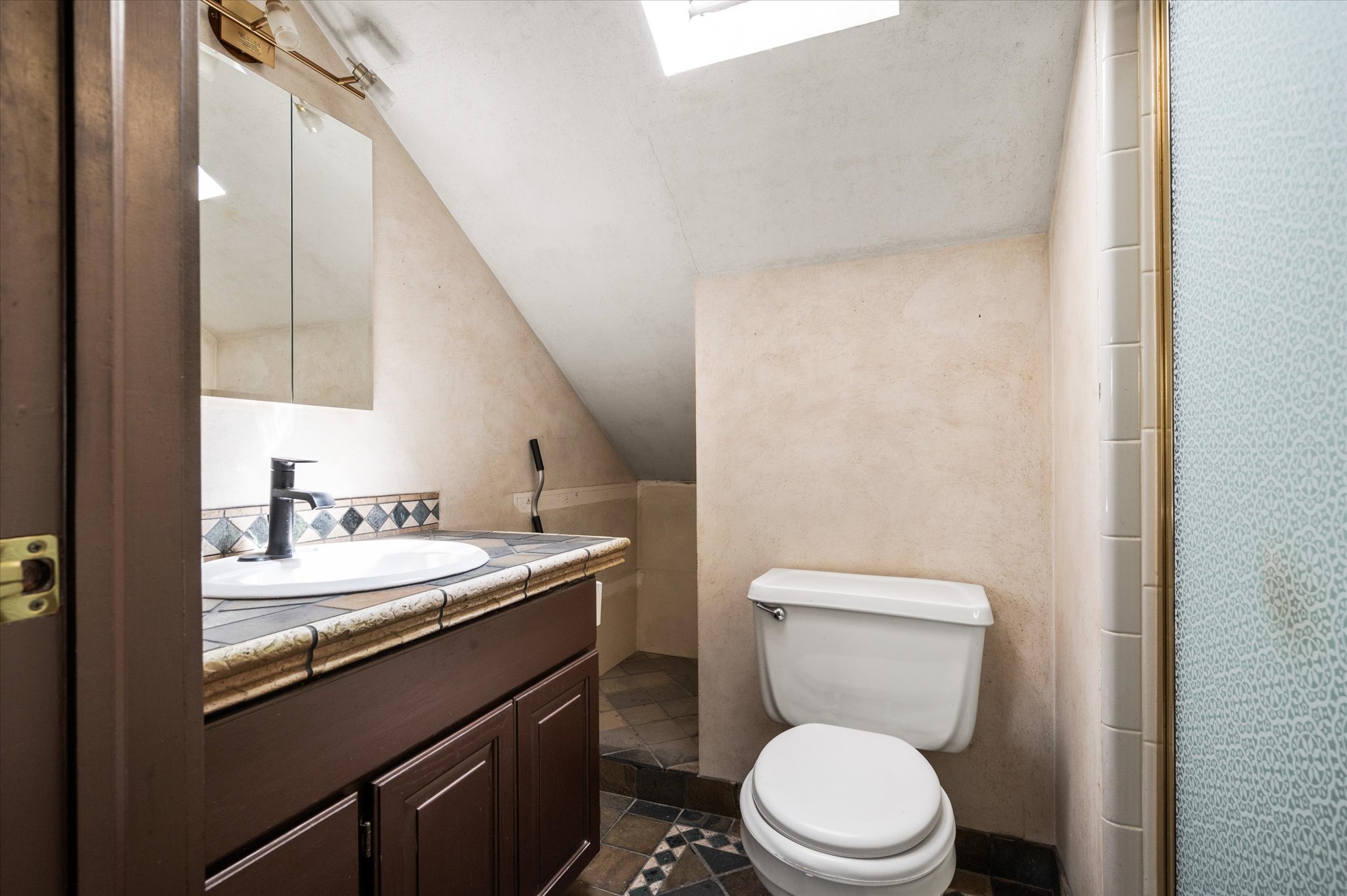 Bathroom featuring a skylight, dark wood vanity with a white basin sink, tiled countertop, and a decorative tile backsplash