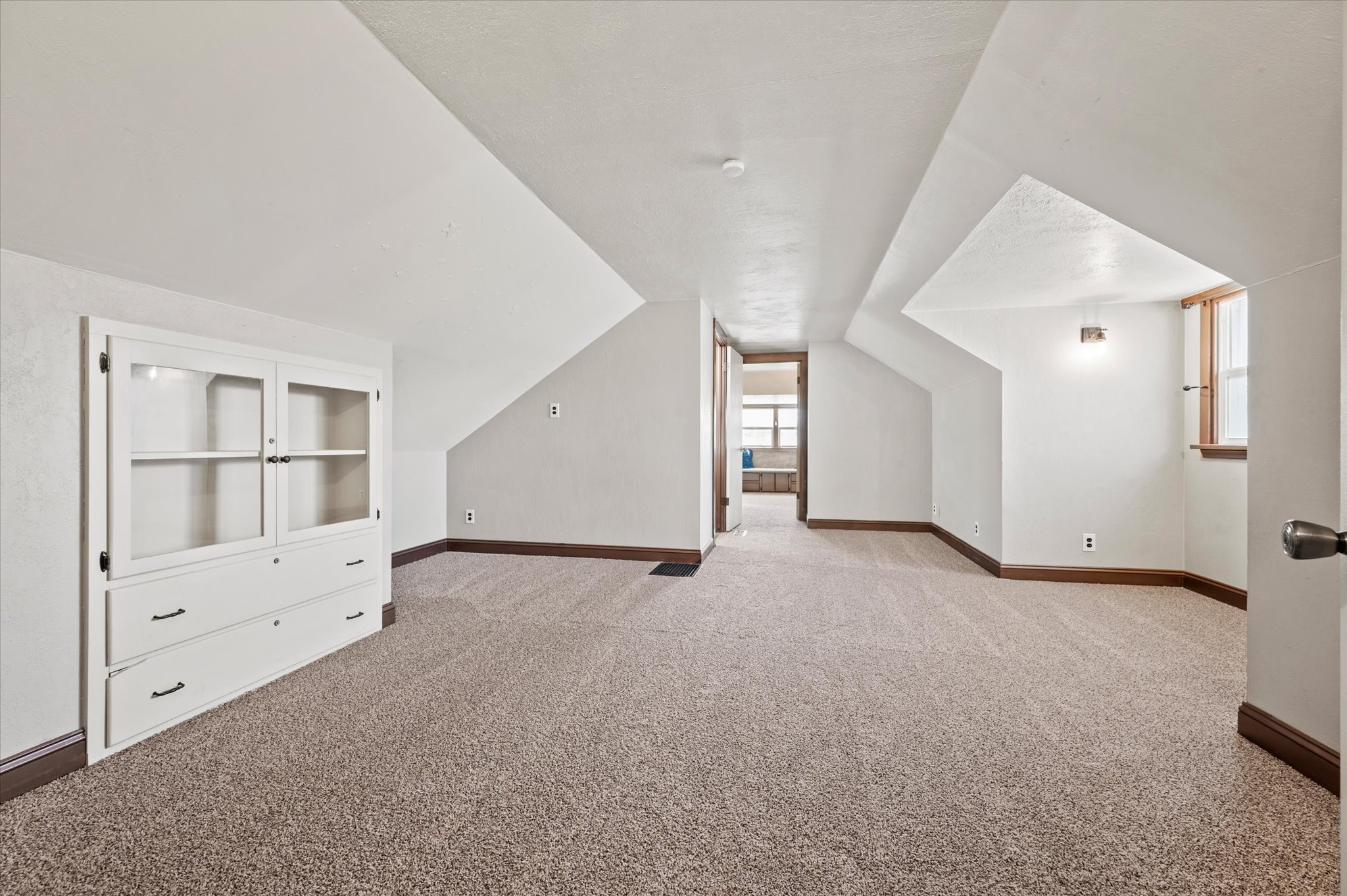 Spacious carpeted room featuring a built-in display cabinet with glass doors and drawers, painted white walls, and dark wood-finish baseboards
