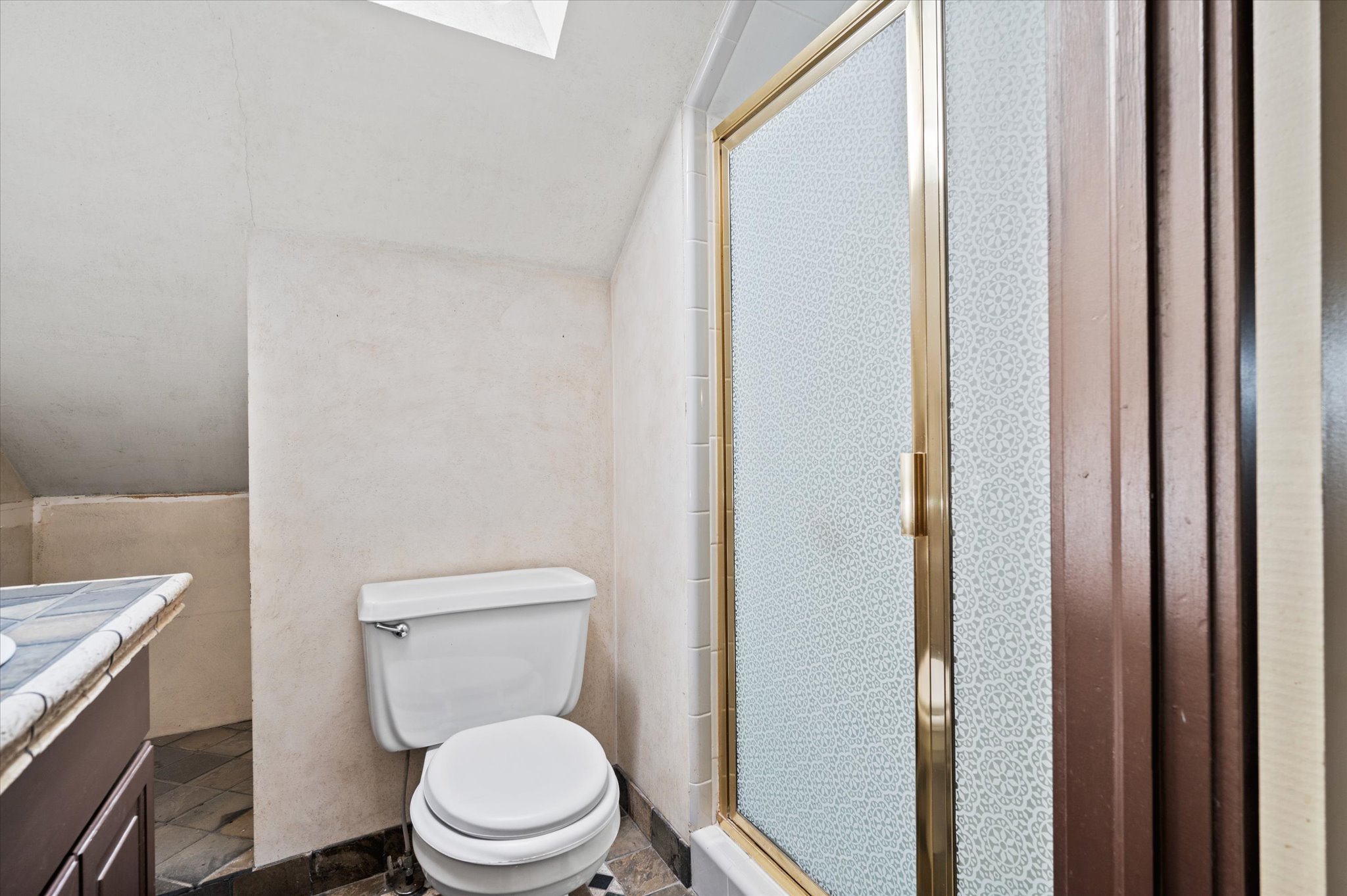 Bathroom featuring a white toilet, a shower with a gold-framed frosted glass door, a vanity with a tiled countertop, and dark-toned flooring