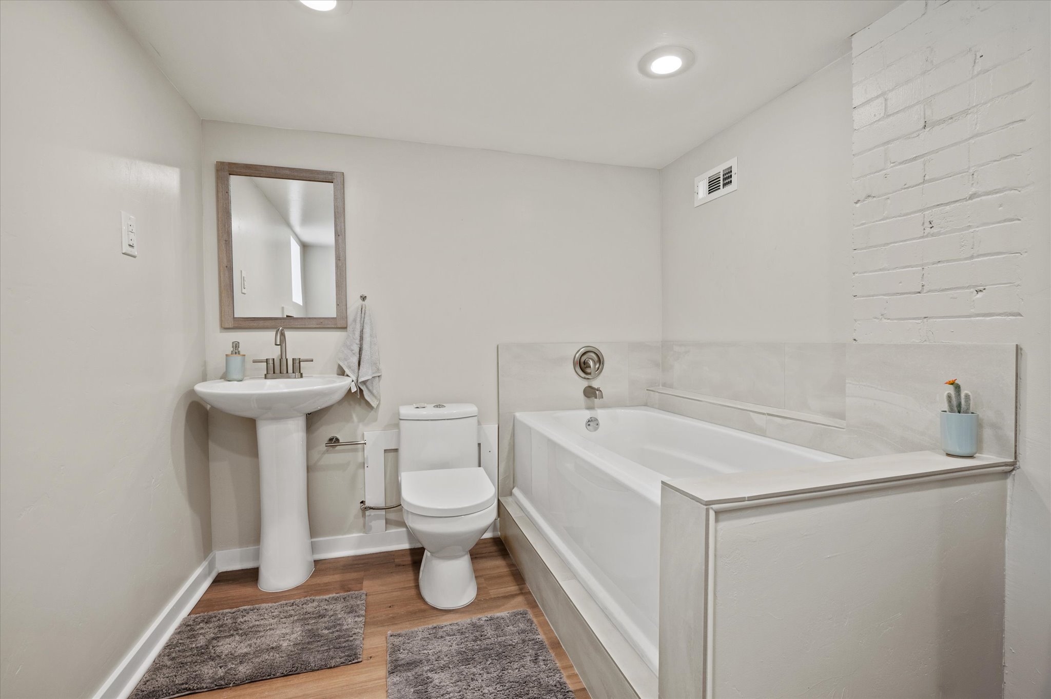 Bathroom featuring a built-in tub with tile surround, pedestal sink, wood-finish flooring, and recessed lighting