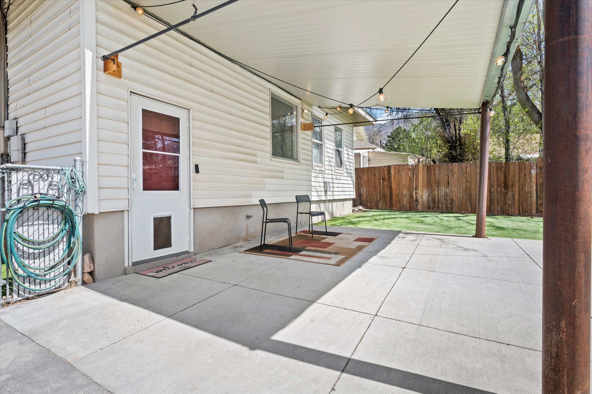 Covered concrete patio with string lighting, white vinyl siding, a rear door with a lower panel, and a wood fence
