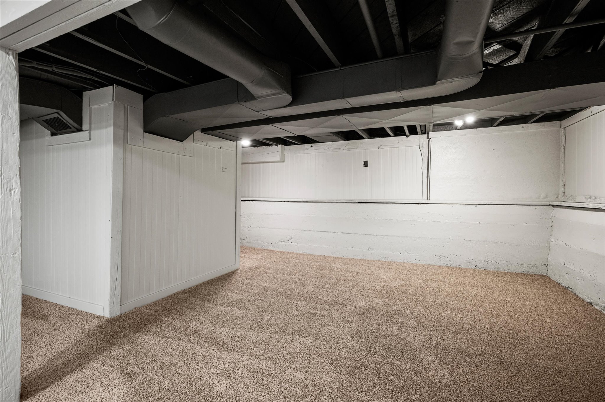 Finished basement space featuring light brown carpet, painted white walls, exposed black ceiling joists, and visible ductwork