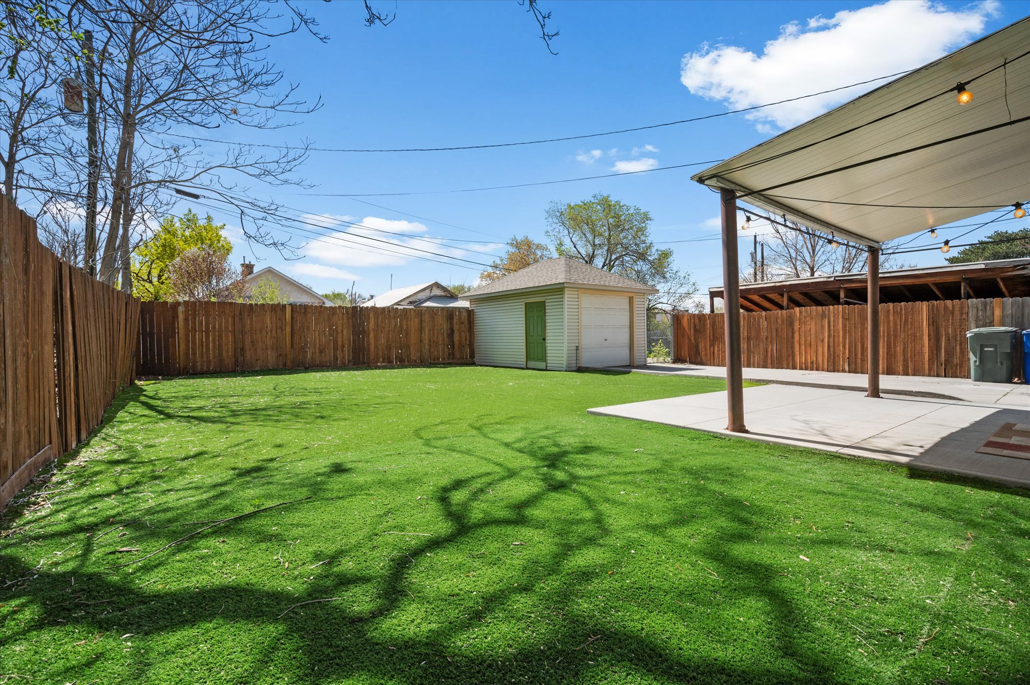 Expansive backyard featuring artificial turf, a privacy fence, a detached utility building, a covered patio, and string lighting