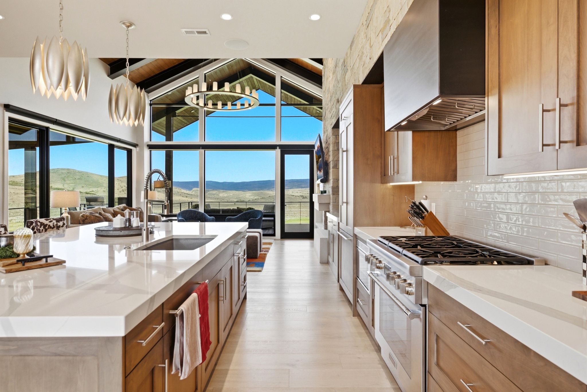Open-concept kitchen featuring an expansive island with an undermount sink, light wood-finish cabinetry, stainless steel appliances, a white subway tile backsplash, and a vaulted ceiling with exposed beams