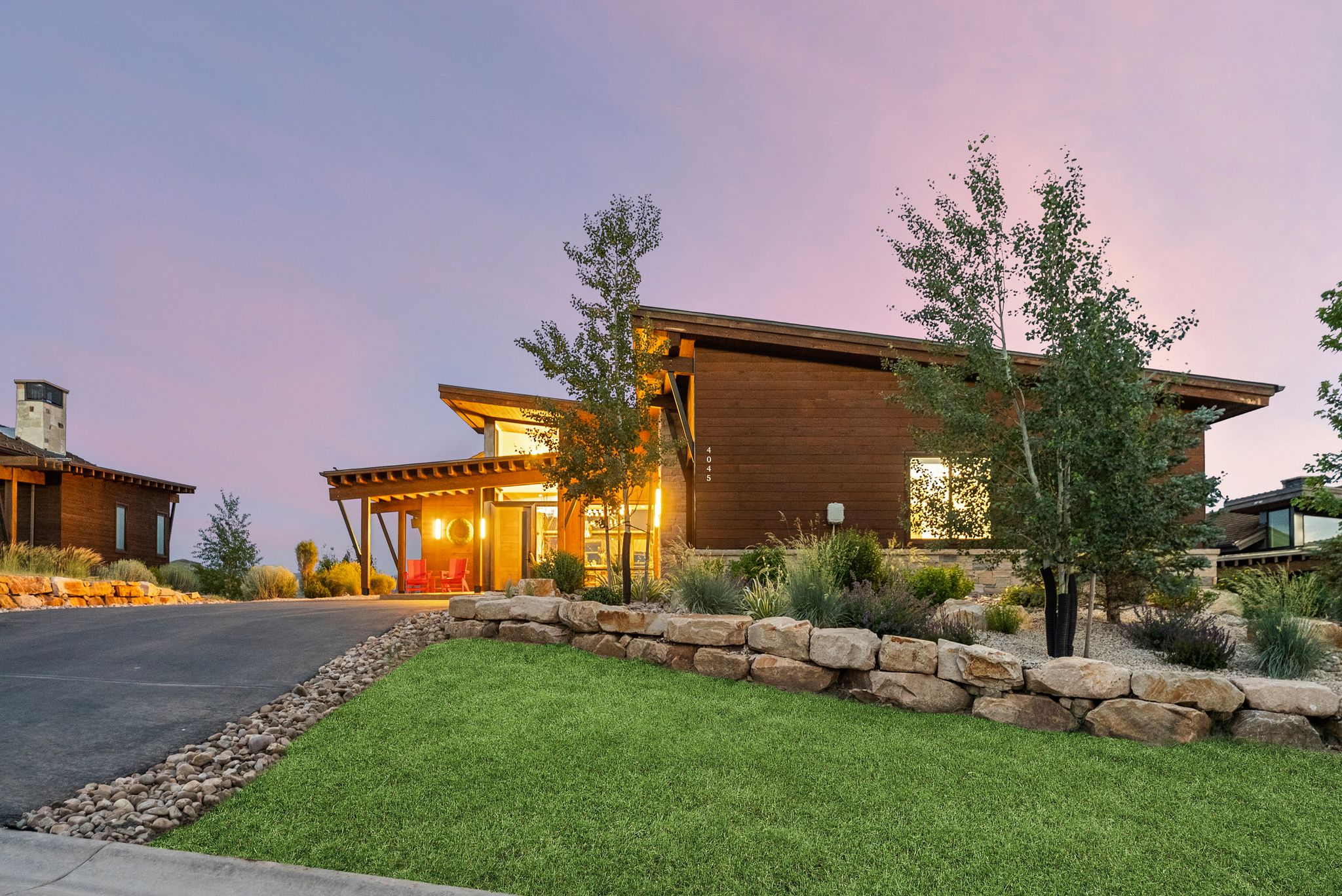 Modern architectural exterior featuring wood siding, an asphalt driveway, a stone retaining wall, and integrated landscaping with mature trees and shrubs