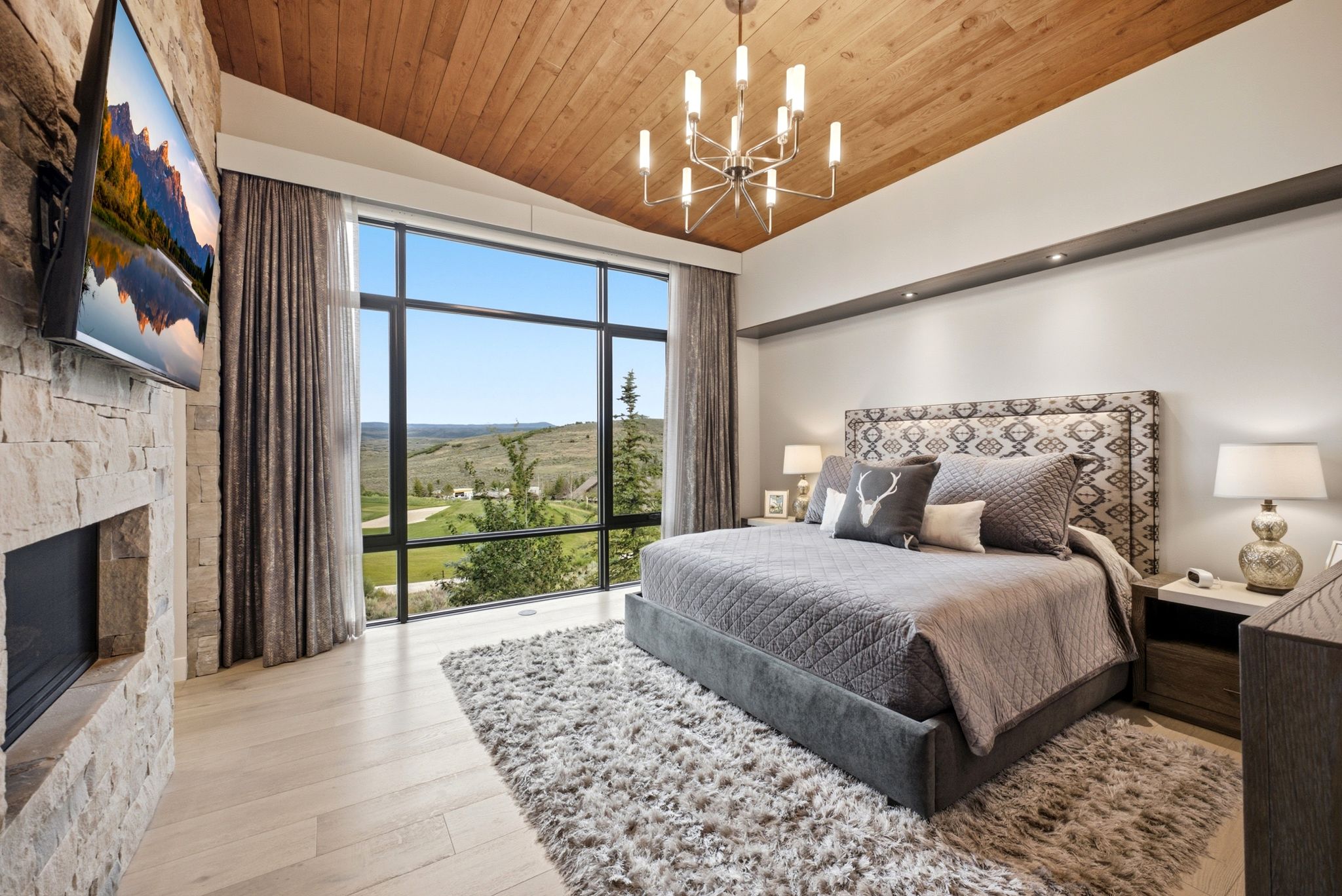 Expansive floor-to-ceiling window overlooking a natural landscape, featuring a stone-clad fireplace, light wood-finish flooring, a wood-paneled vaulted ceiling, and a contemporary chandelier