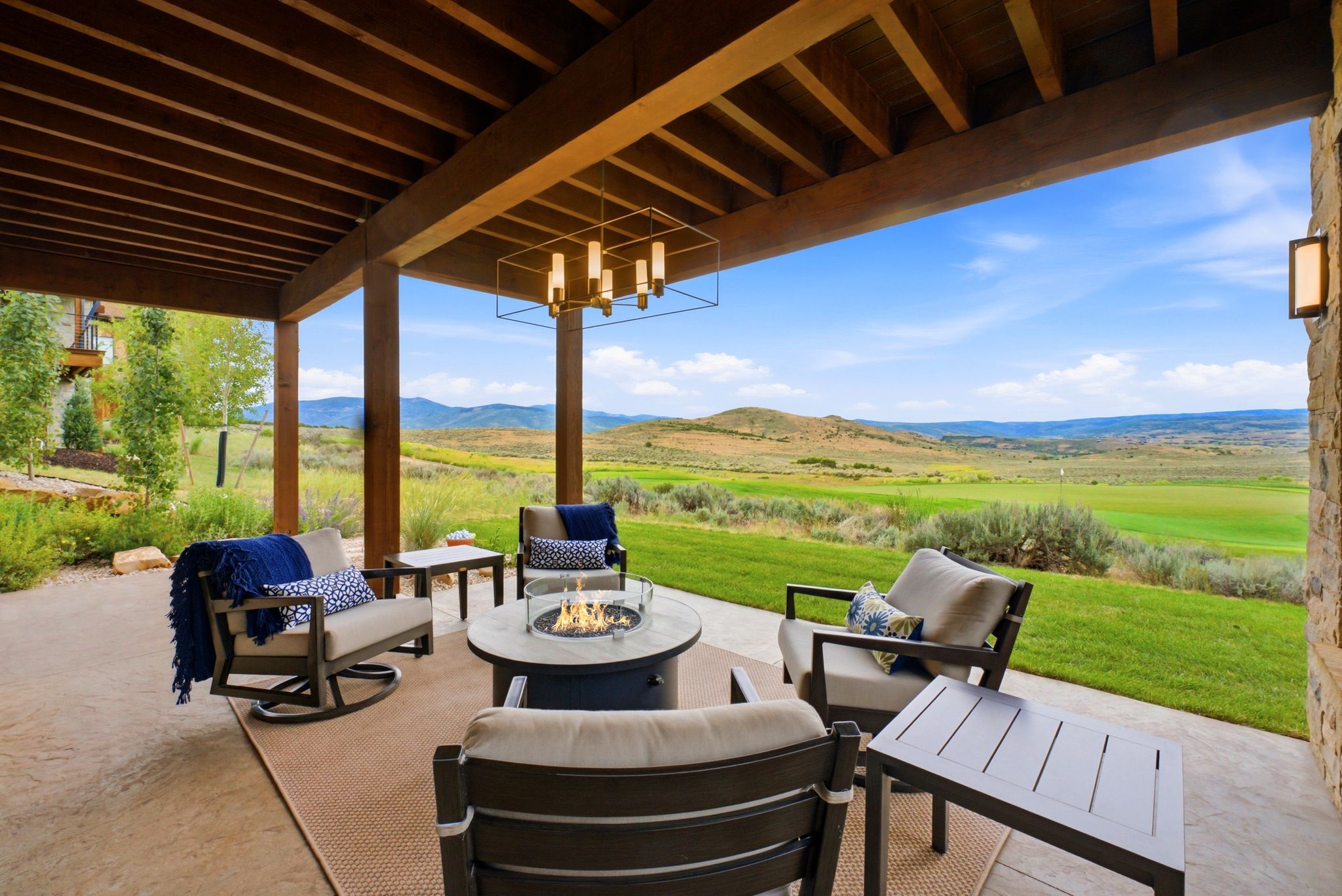Covered patio featuring an exposed wood beam ceiling, contemporary chandelier, stone accent wall, and a built-in fire pit