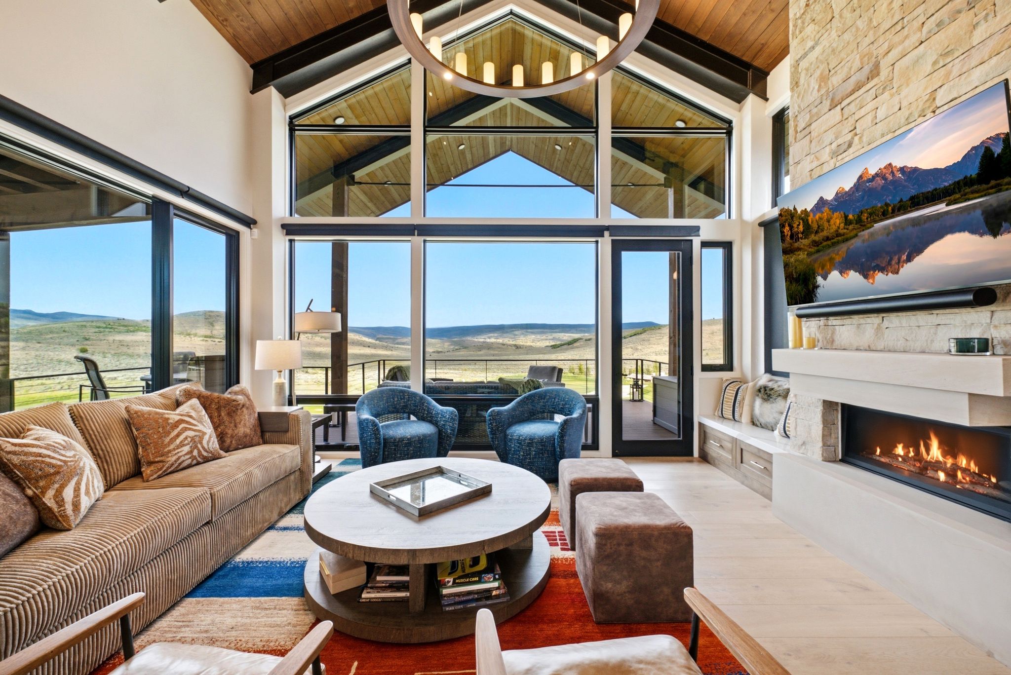Vaulted wood-paneled ceiling with exposed beams, expansive window wall, light wood-finish flooring, linear fireplace with stone surround, and built-in bench seating