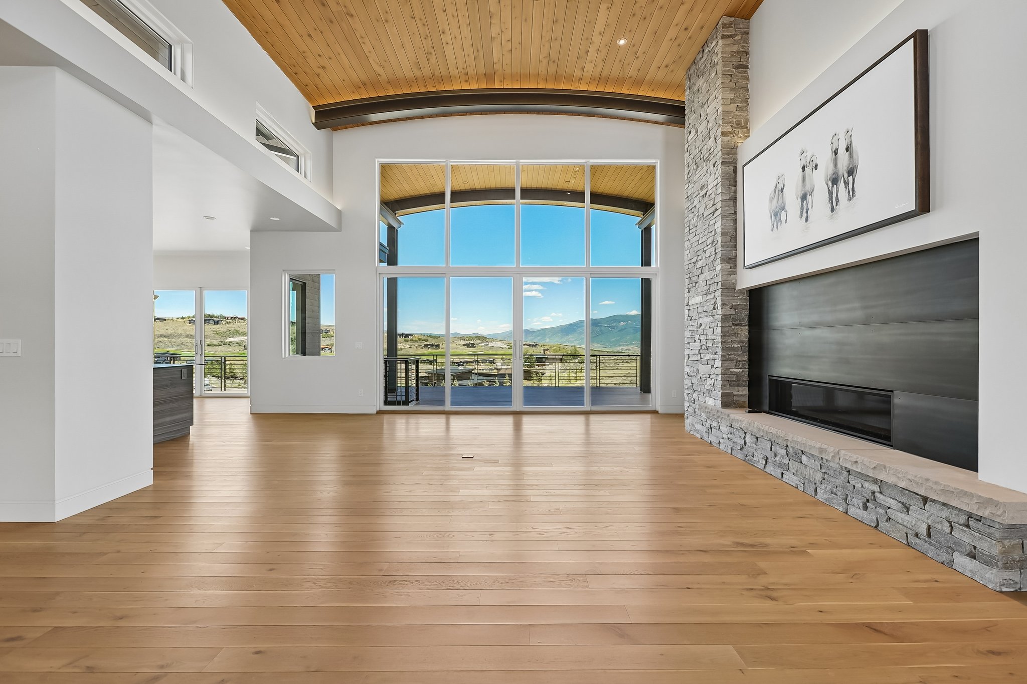 Expansive living space featuring a floor-to-ceiling window wall with mountain scenery, a stone-clad fireplace with a dark-toned hearth, warm wood-finish flooring, a wood-paneled vaulted ceiling, and recessed lighting