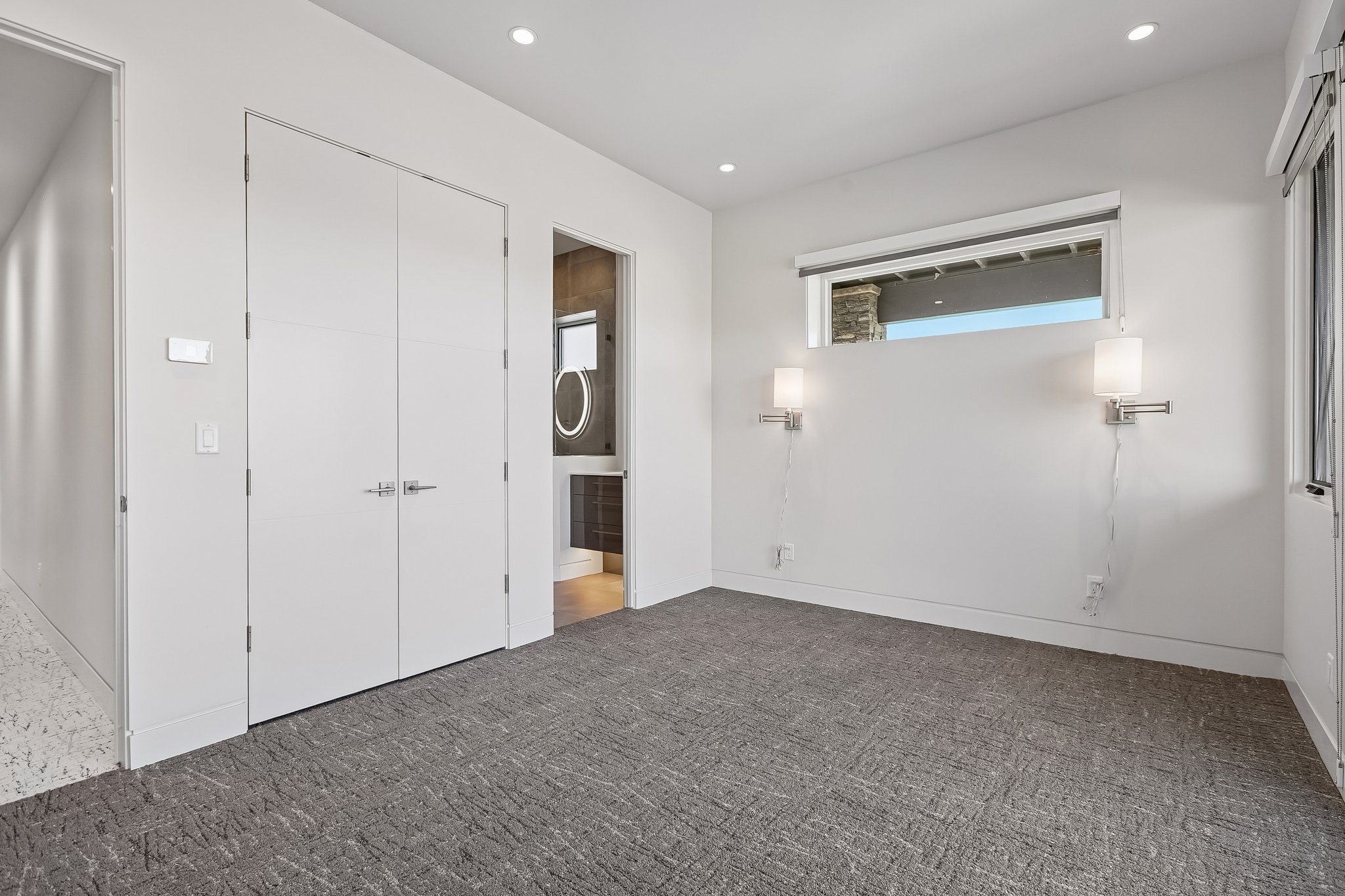 Spacious room featuring patterned gray carpeting, dual white doors, a high-set horizontal window, and two wall-mounted sconces