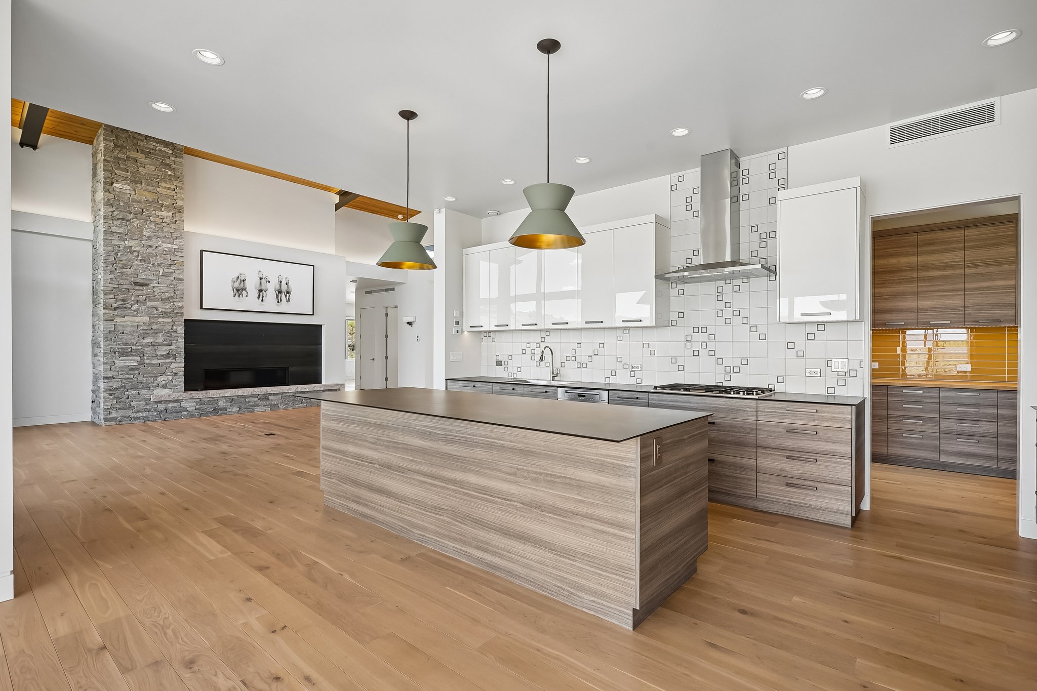 Expansive open-concept space featuring wood-finish flooring, a stone-clad fireplace, a large kitchen island with a dark countertop, white upper cabinetry, and stainless steel kitchen appliances