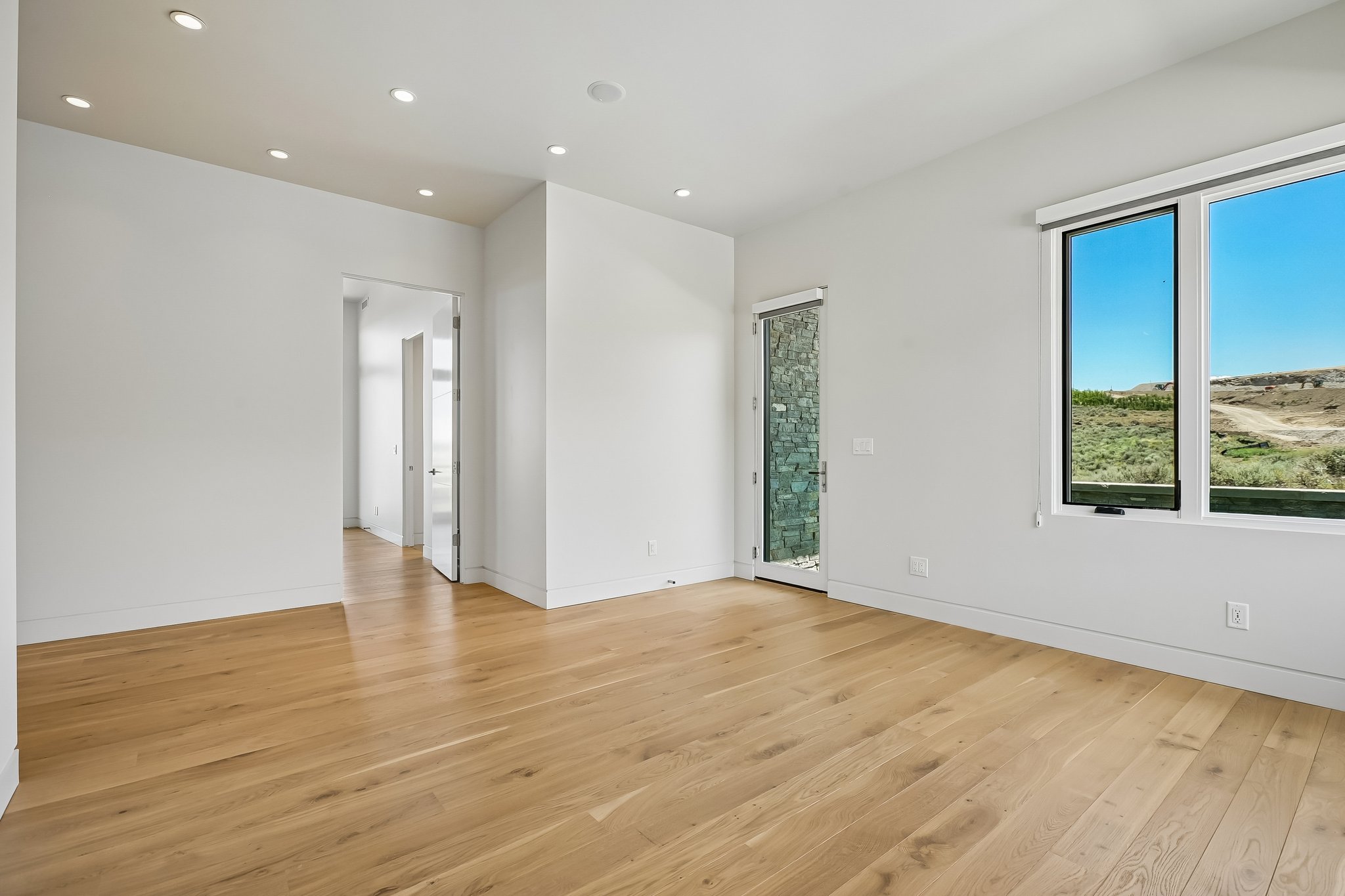 Spacious room featuring wood-finish flooring, recessed ceiling lighting, a door with stone-look exterior, and expansive windows