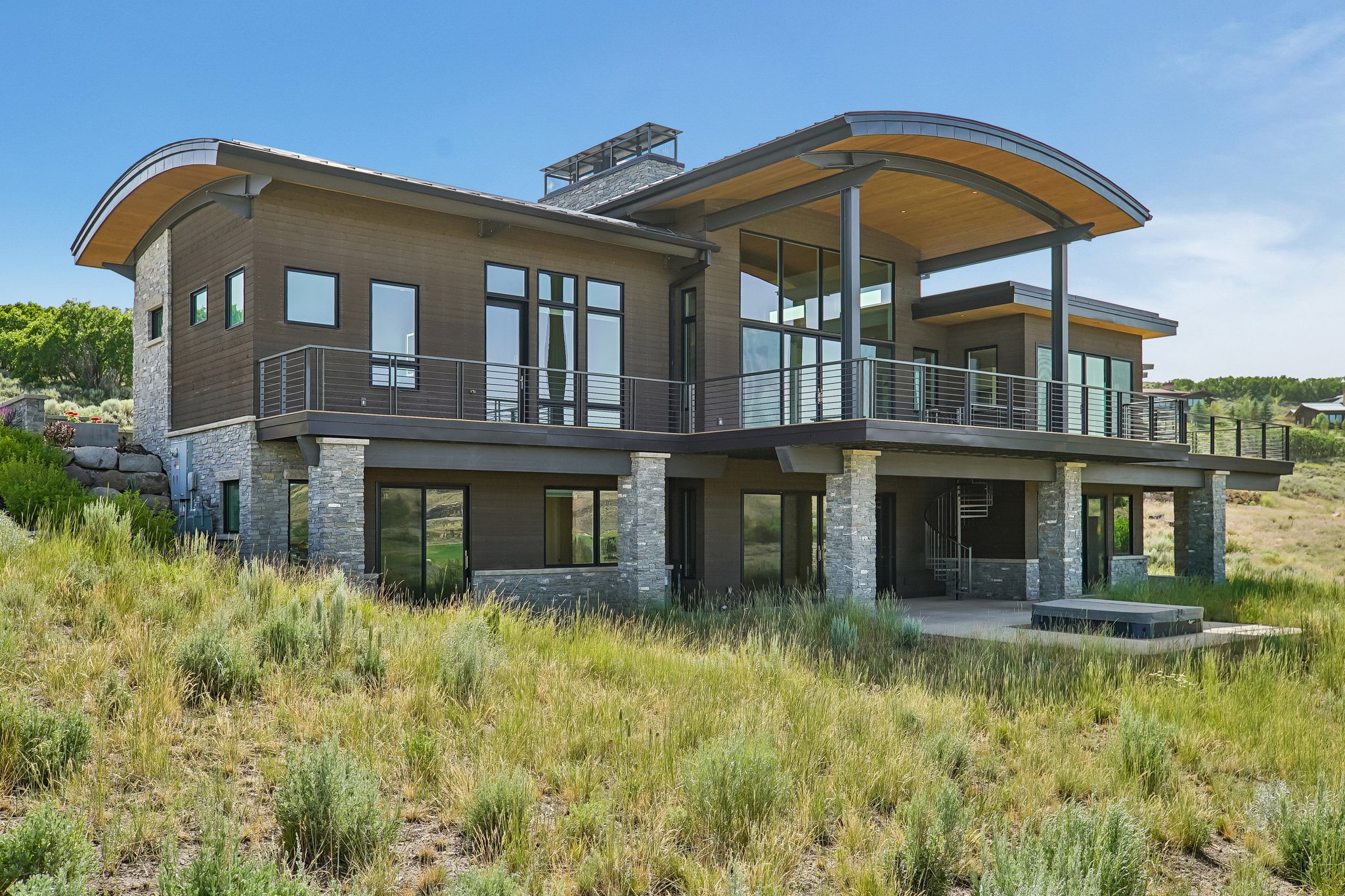 Contemporary architecture featuring a curved wood-paneled soffit, extensive glass windows, multiple balconies with cable railings, stone columns, and a lower-level patio with a built-in fire feature