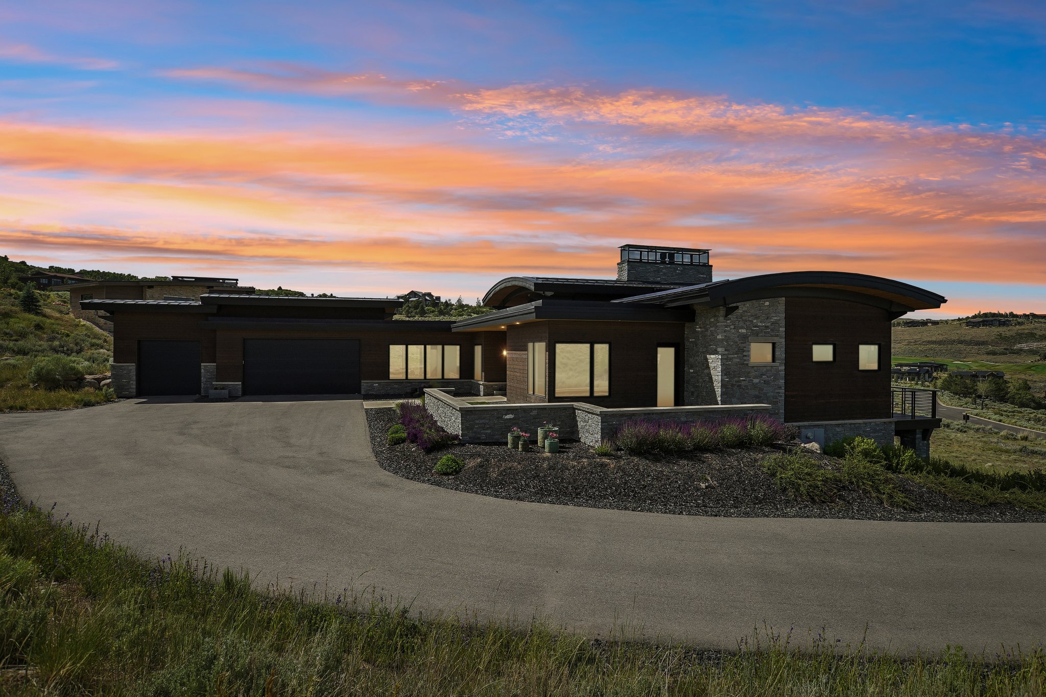 Contemporary architecture featuring a curved roofline, dark wood siding, stone accents, multiple large windows, and an expansive driveway