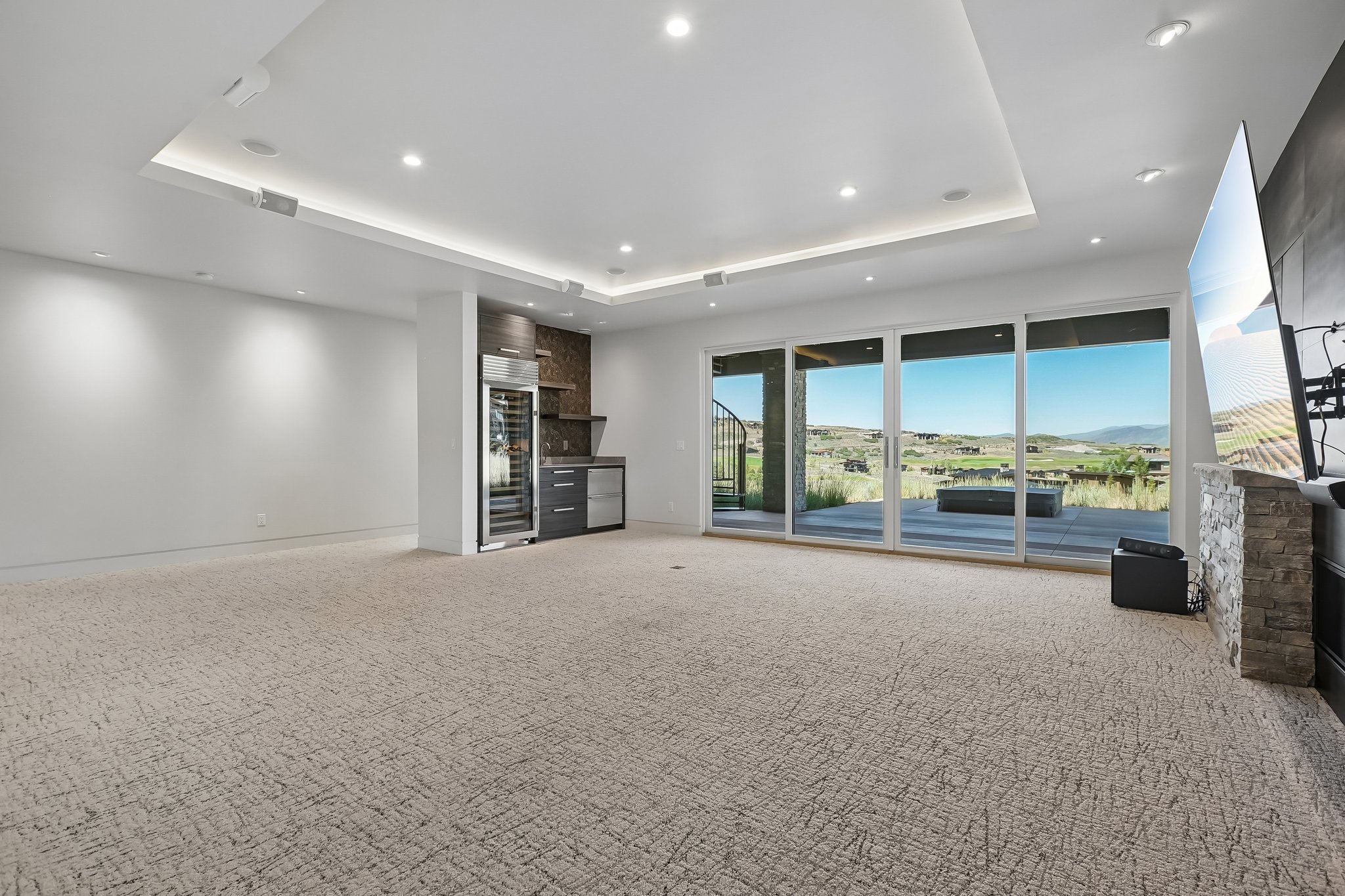 Expansive room featuring multi-panel sliding glass doors, a built-in wet bar with a wine refrigerator, a stone-clad fireplace, and recessed ceiling lighting