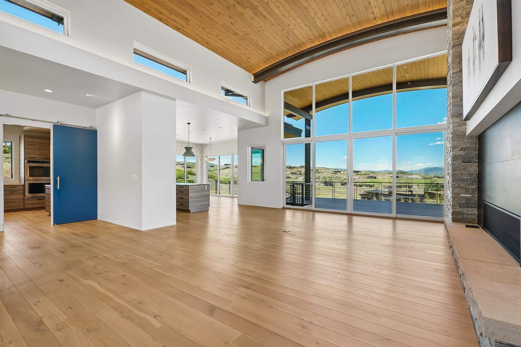 Spacious living area featuring expansive floor-to-ceiling windows, wood-finish flooring, a wood-paneled vaulted ceiling, a stone-clad fireplace surround, and a blue barn door leading to a kitchen with wood cabinetry