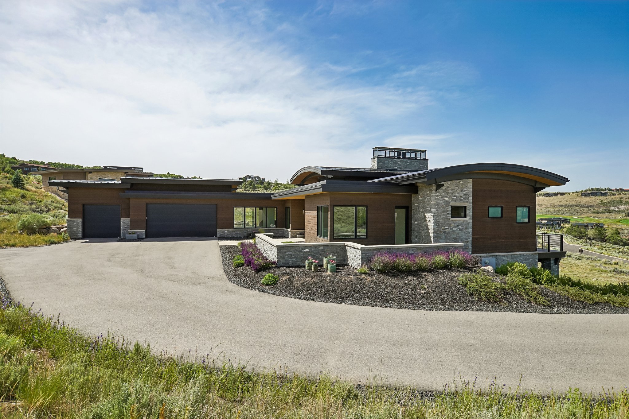 Modern architectural residence featuring a curved roofline, stone and wood-paneled exterior, multiple large windows, a two-car garage, and a winding driveway