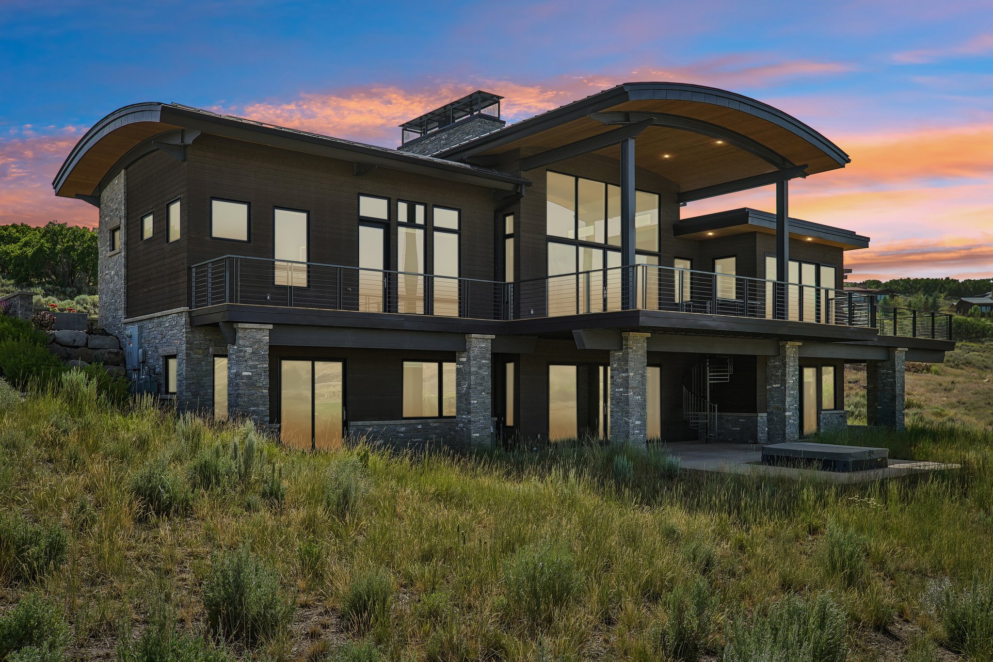 Modern architectural design featuring extensive glazing, a curved roofline, stone column accents, multiple balconies with cable railings, and a lower-level patio
