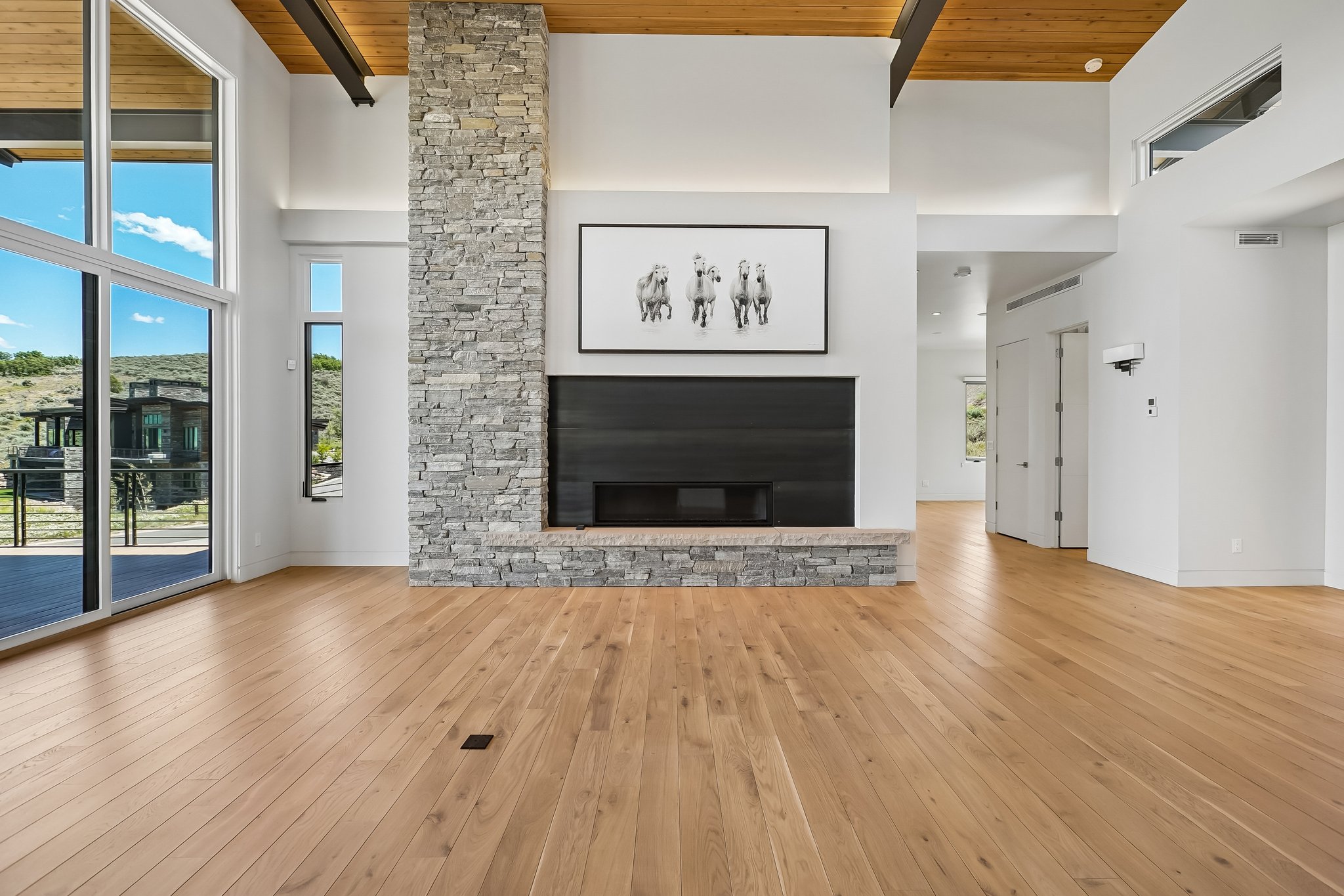 Spacious living area featuring a floor-to-ceiling stone fireplace, wood-finish flooring, high ceilings with exposed wood beams, and expansive glass sliding doors opening to an exterior deck