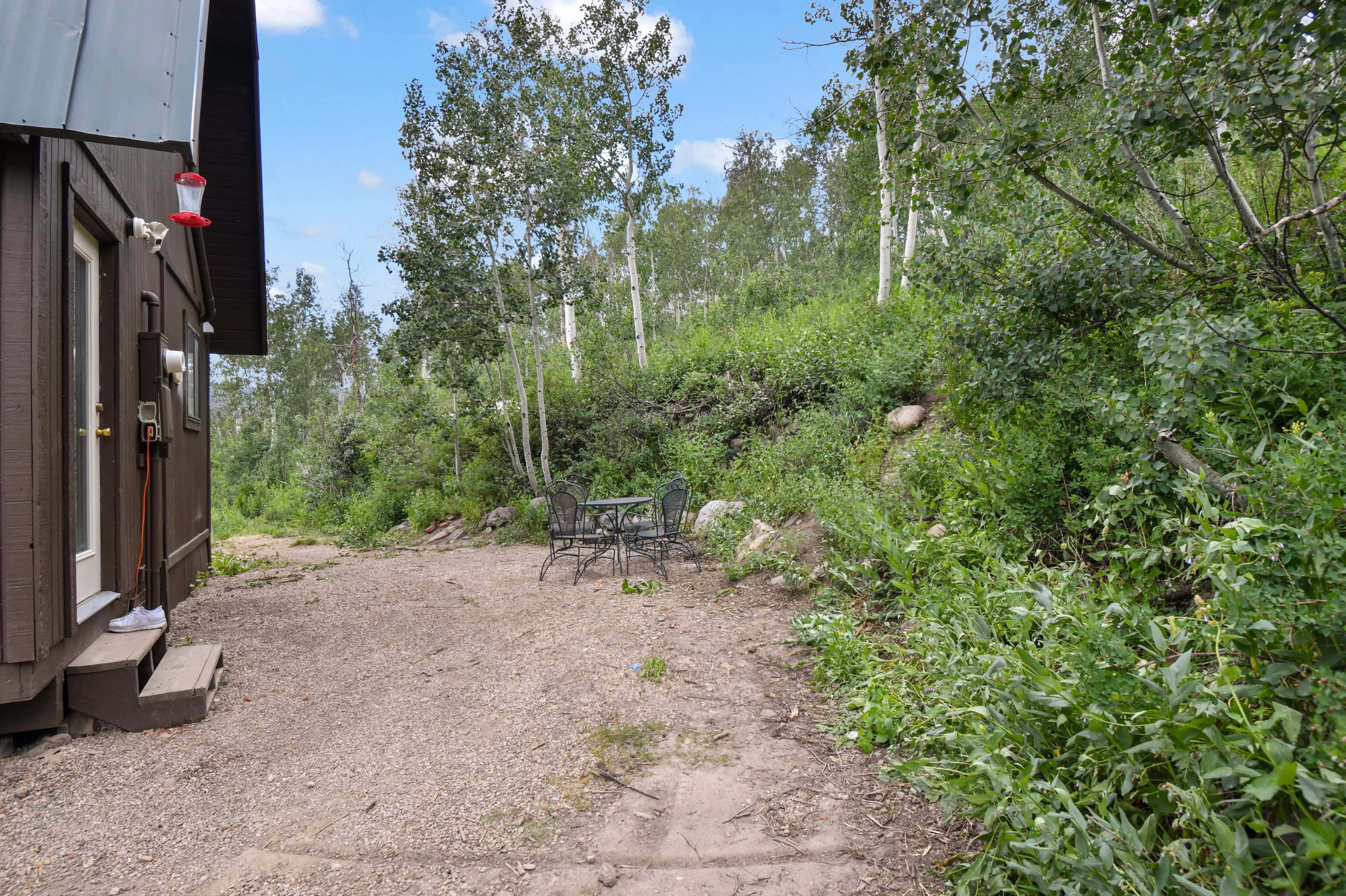 Gravel patio area featuring a metal outdoor dining set, surrounded by mature trees and natural landscaping