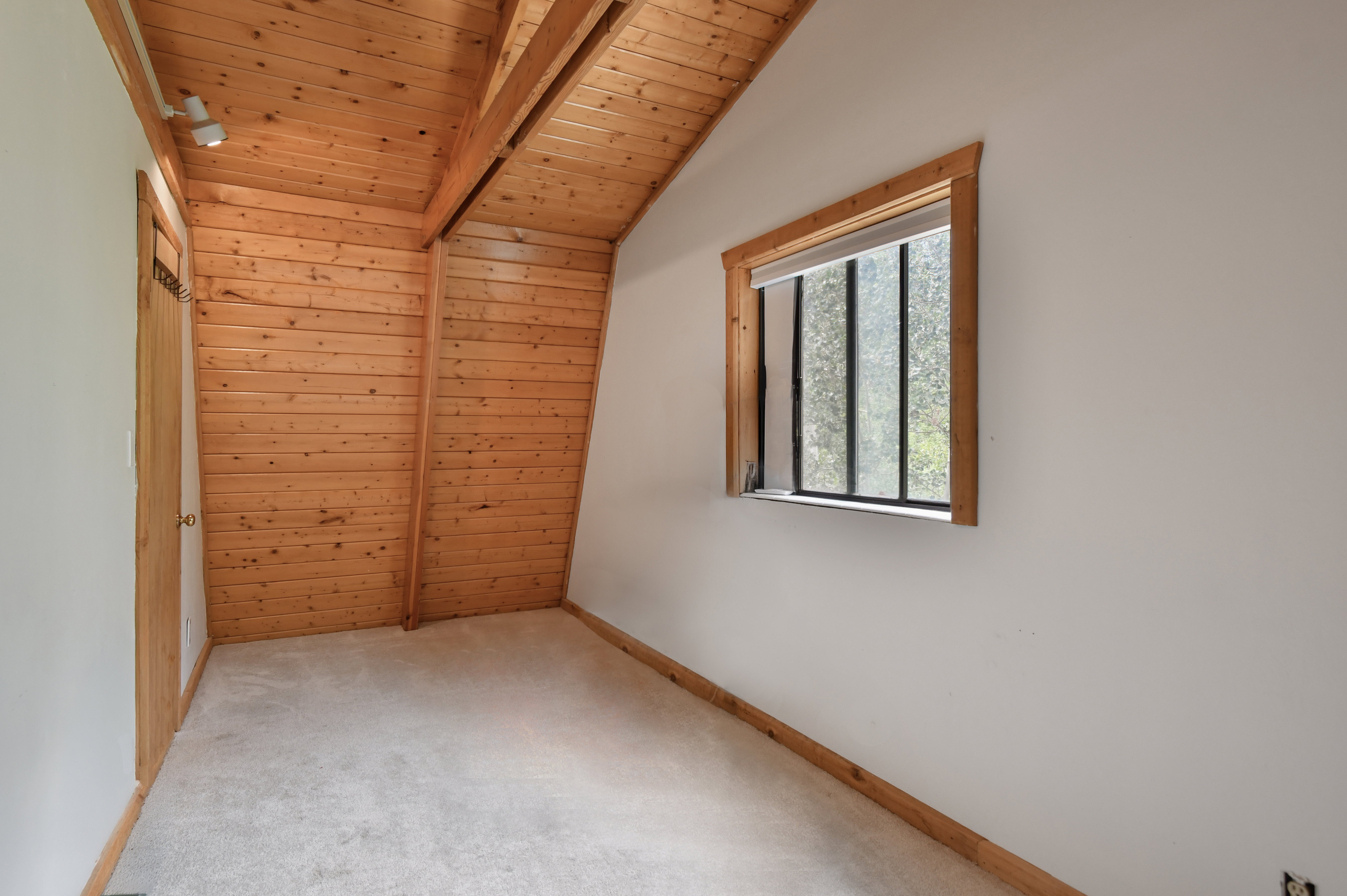 Room featuring an angled wood-paneled ceiling, exposed wood beams, and a window with wood trim