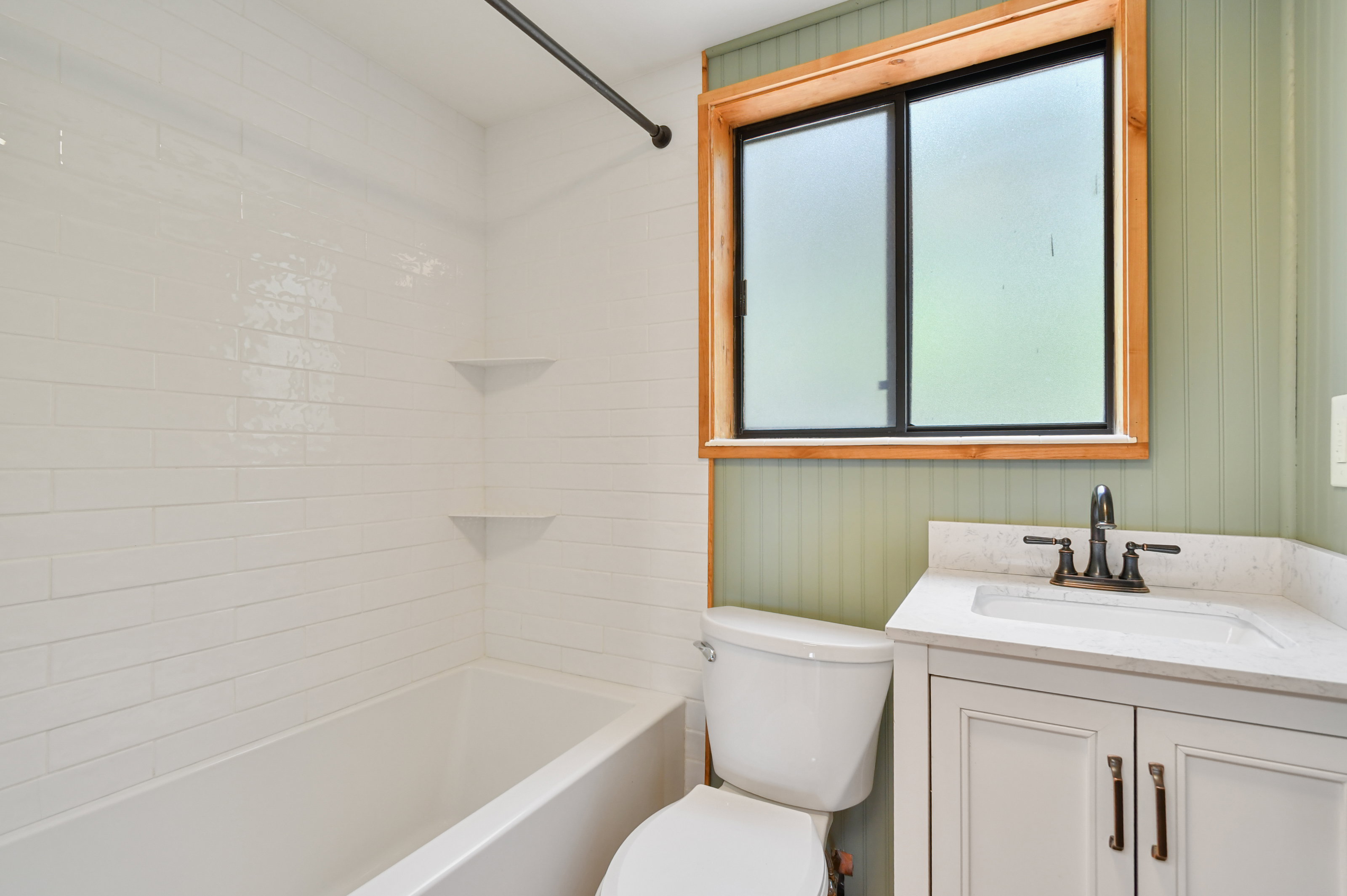 Bathroom featuring a white subway tile tub surround with integrated corner shelves, a natural wood-framed window, and a light green beadboard accent wall