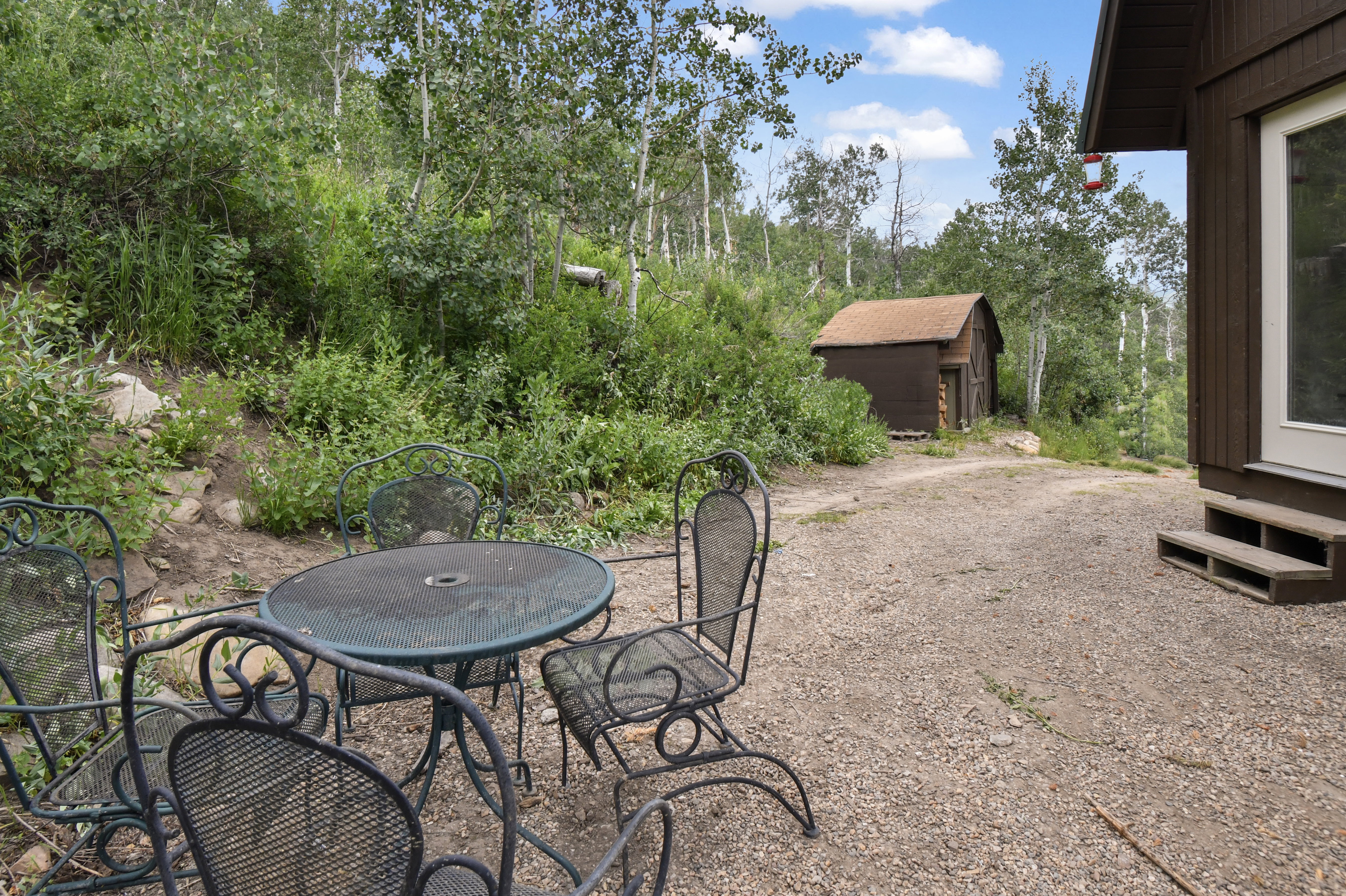 Gravel patio area featuring a detached storage shed, lush natural landscaping, and mature trees
