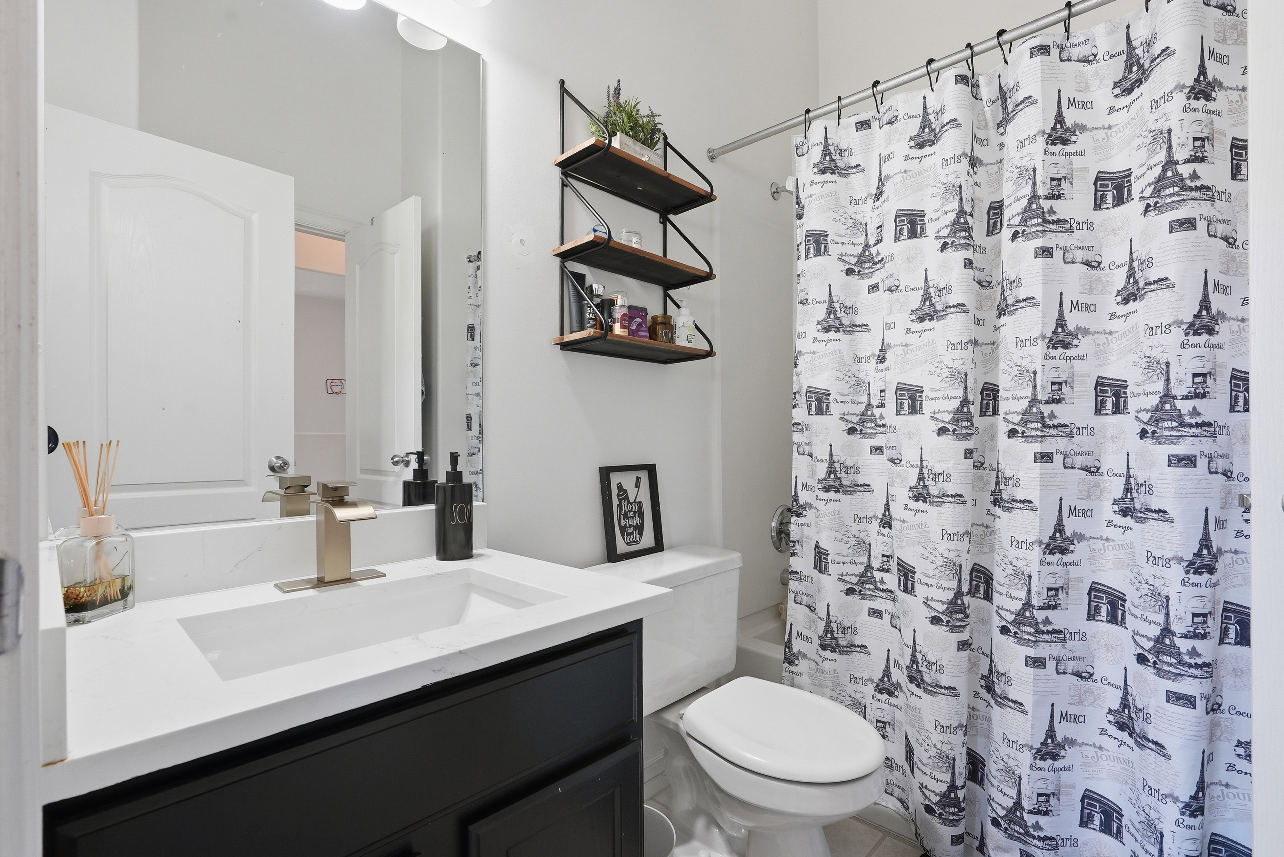 Contemporary bathroom featuring a black vanity with a white countertop and integrated sink, a gold-tone faucet, a wall-mounted mirror, and tiered shelving
