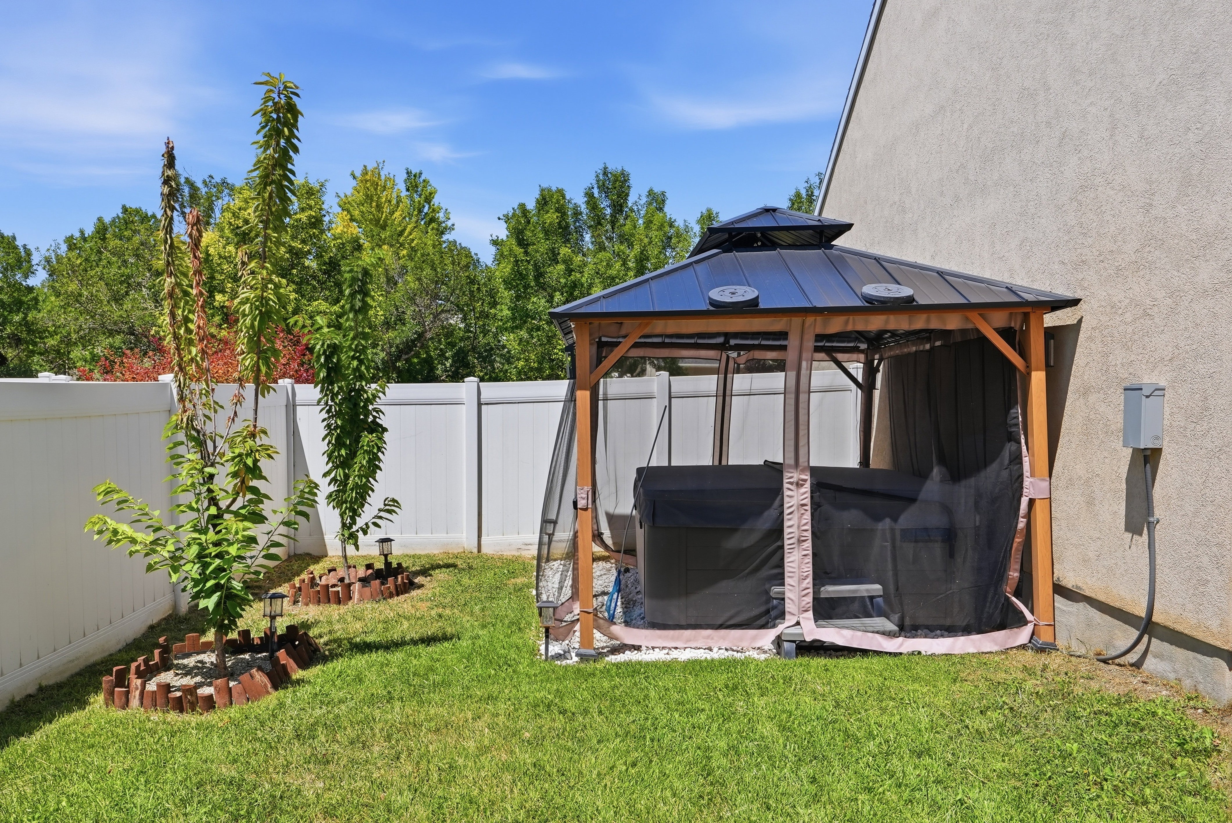 Private outdoor space featuring a covered hot tub, manicured lawn, young trees, and a white privacy fence